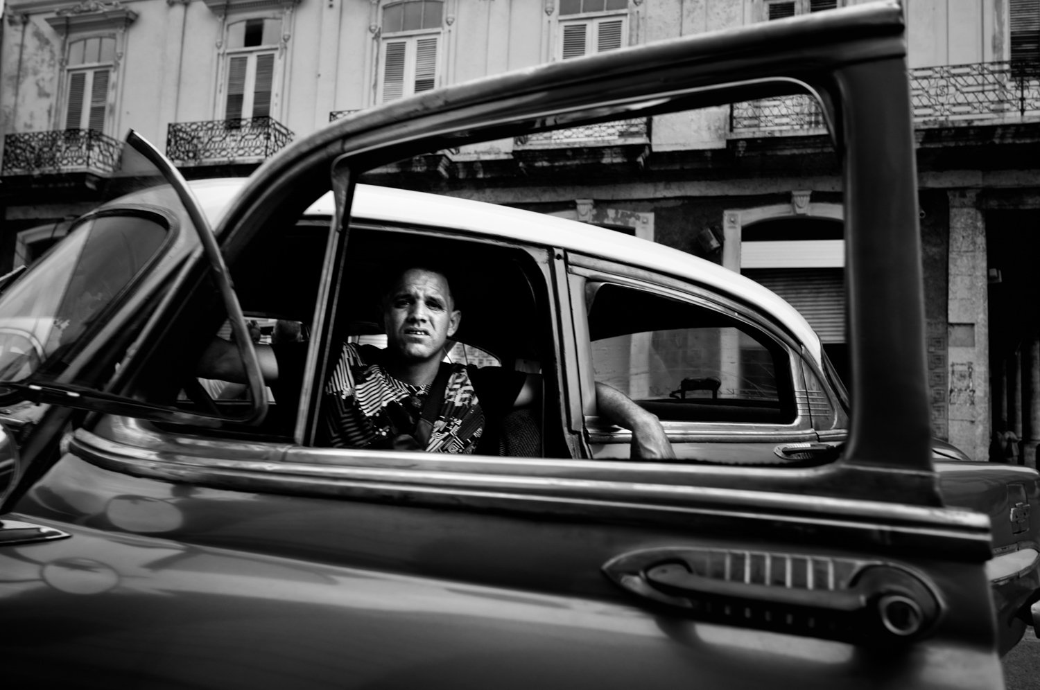 A man looks out from the window of a vintage car parked on a city street, with an old building in the background.