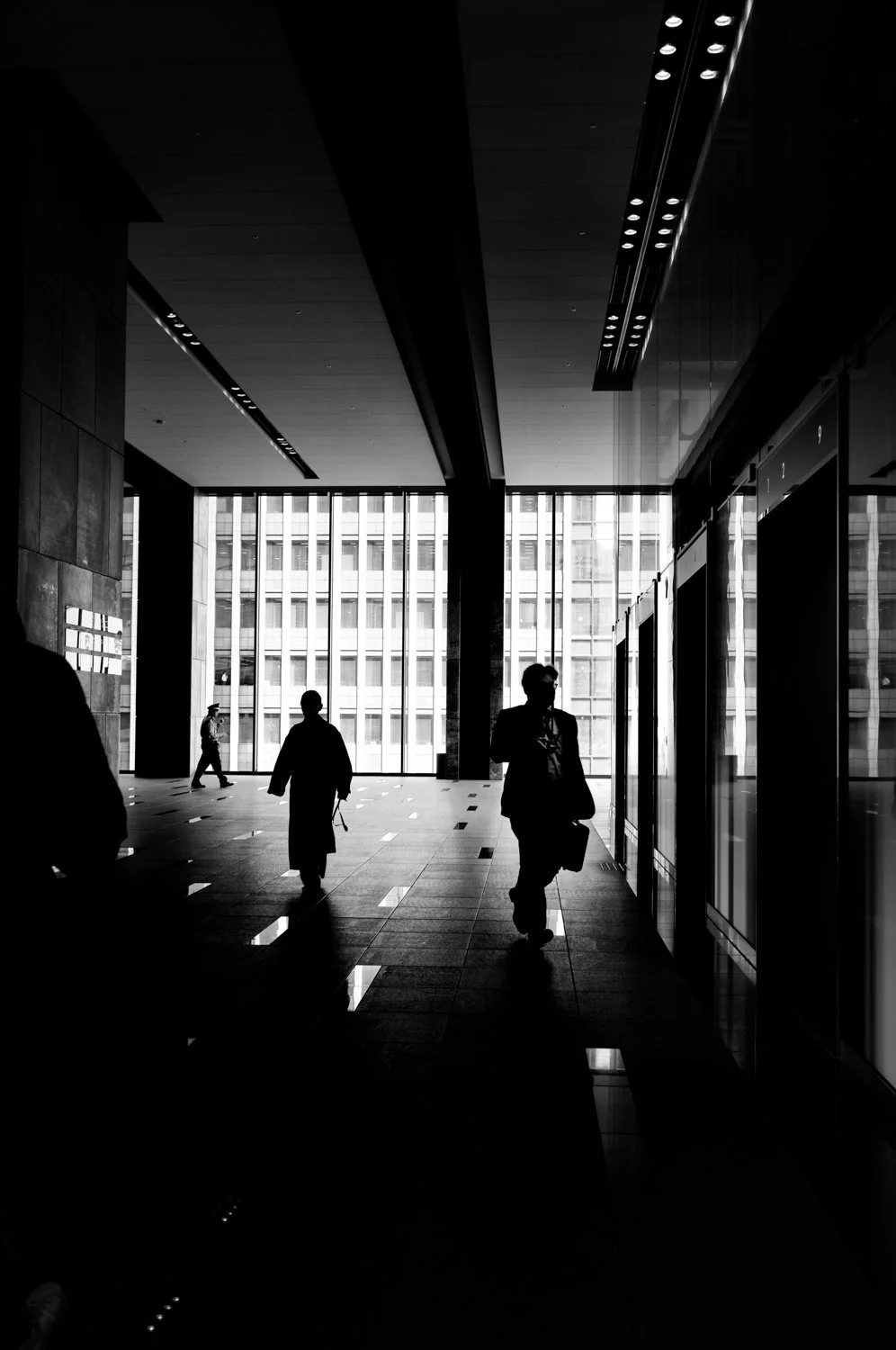 Silhouettes of people walking inside a modern office building with large windows and high ceilings, black and white photograph.