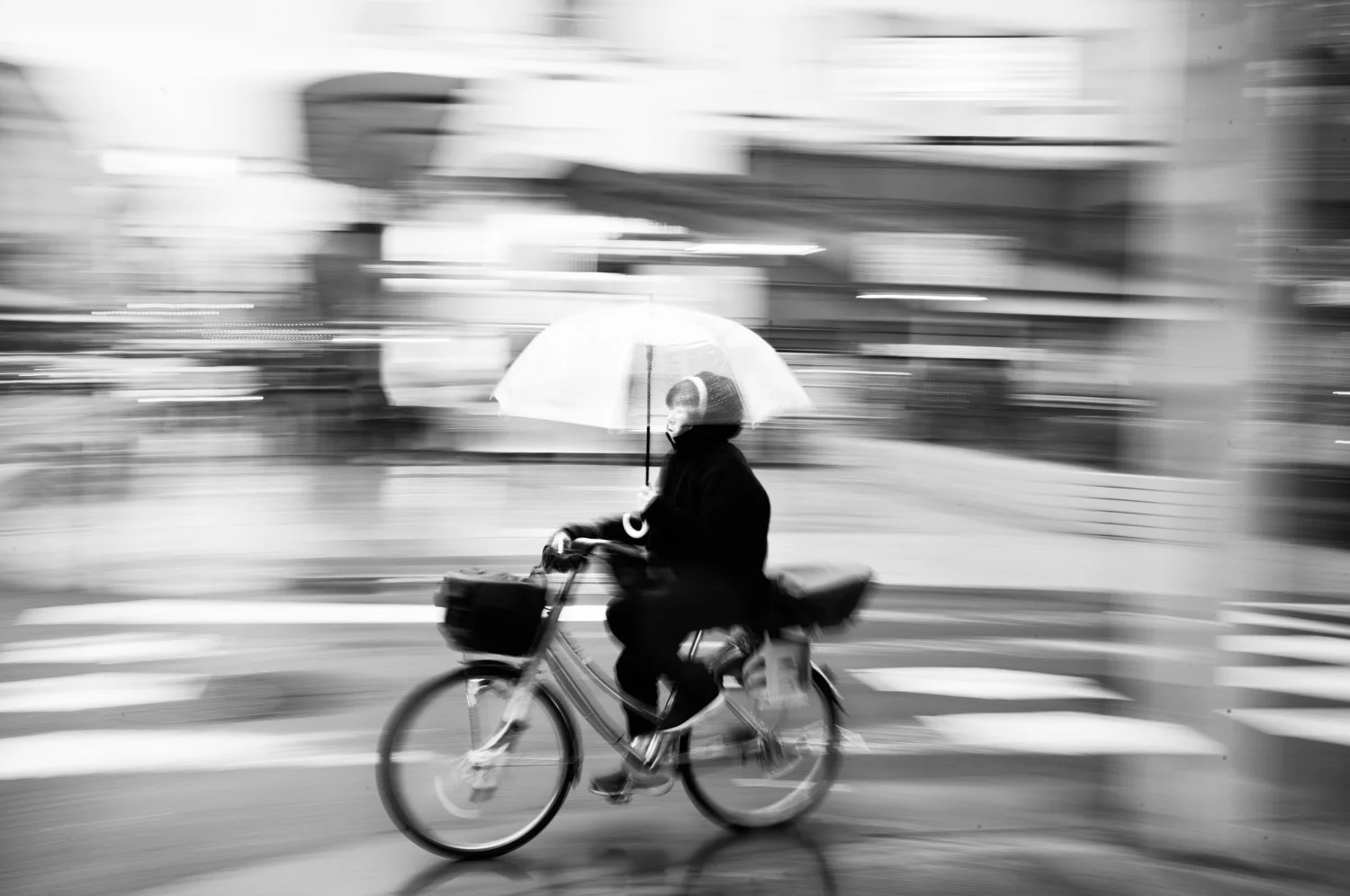 A person riding a bicycle in the rain, holding an umbrella, with a blurred city background in black and white.