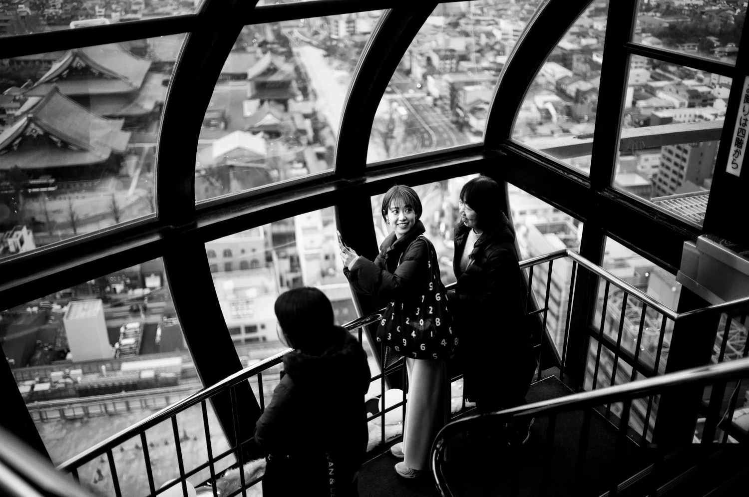 Three women standing on a glass observation deck high above a city, with large windows showing buildings and streets below, one woman smiling at another while taking a photo.