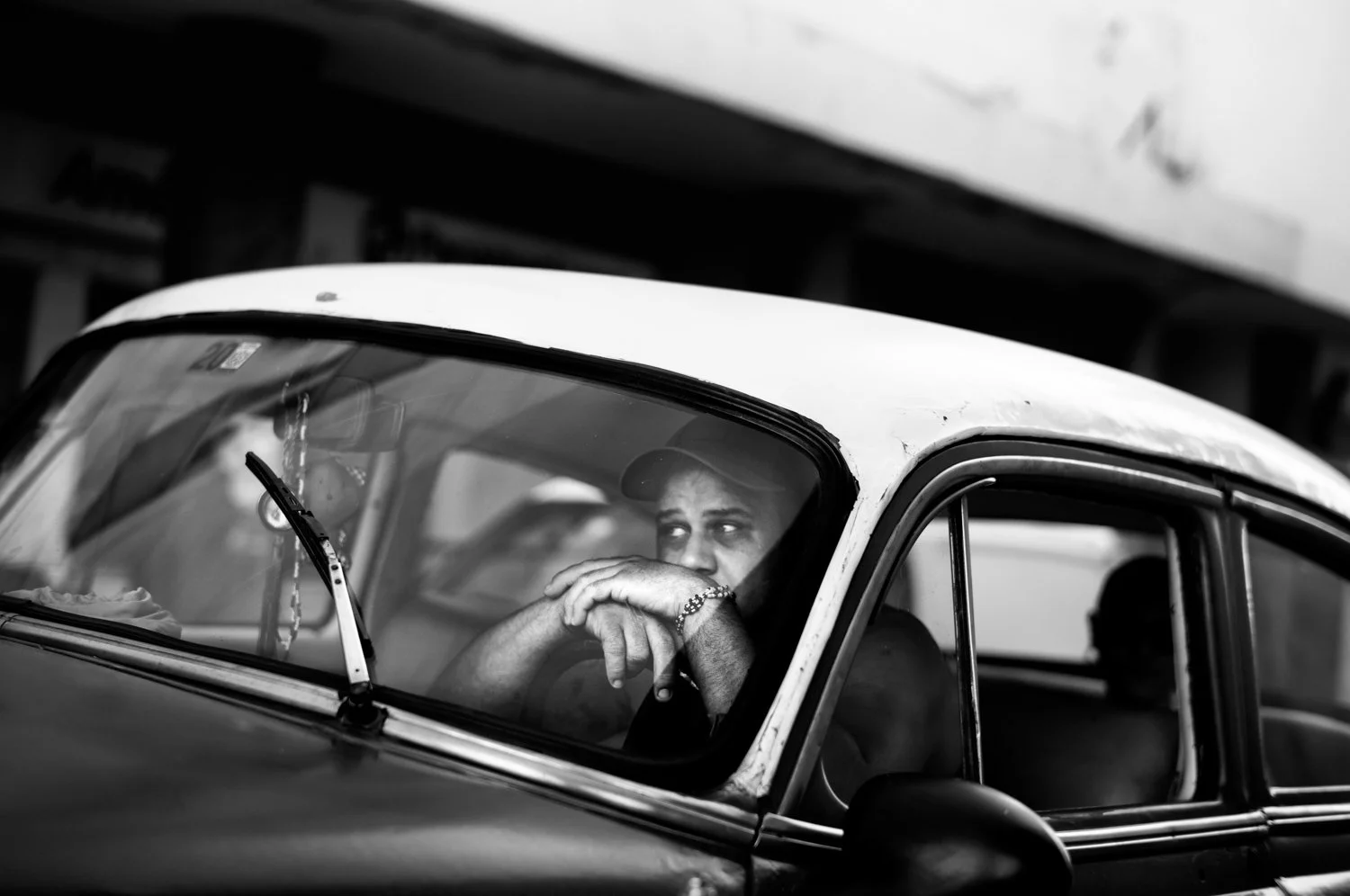 A person sits inside a vintage car, resting their hands on the steering wheel and looking through the front window, in a black-and-white photo.