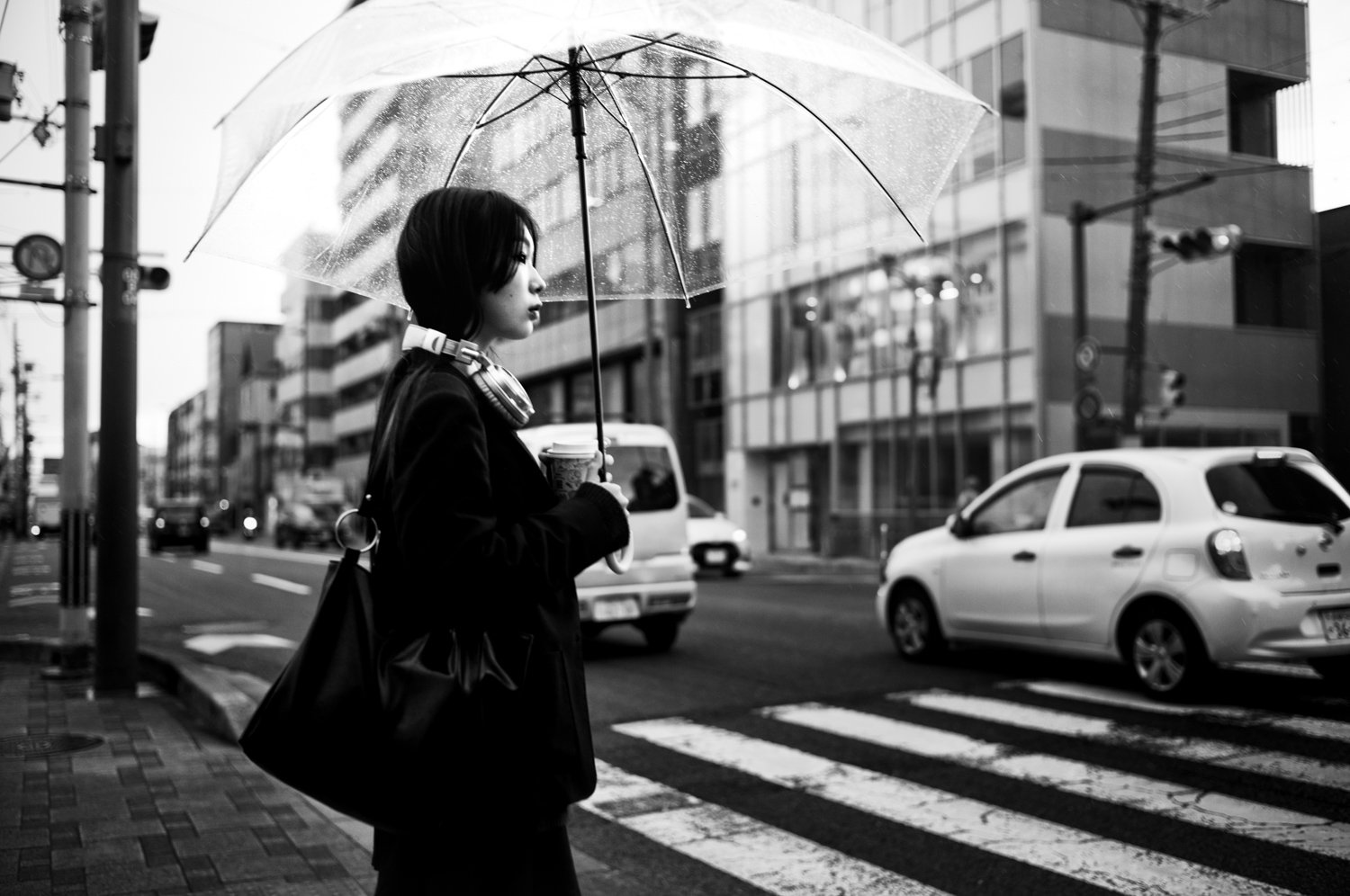 A woman standing at a crosswalk holding a coffee cup and an umbrella during rainy weather in an urban setting.