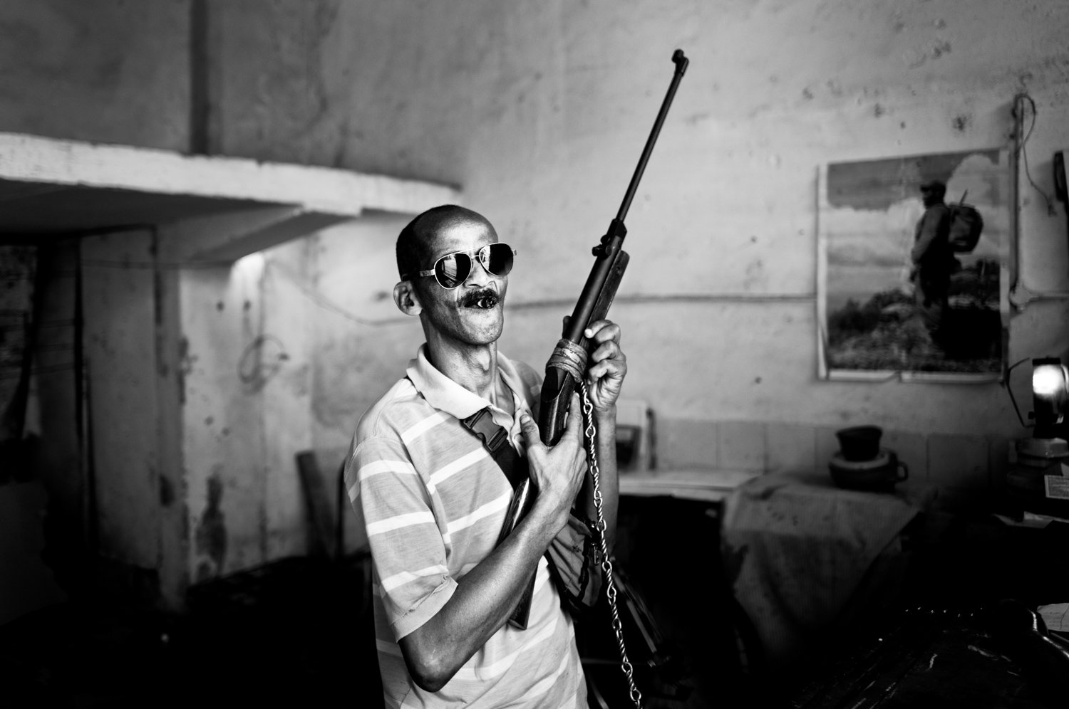 Black and white photo of a man wearing sunglasses, a striped shirt, and holding a rifle inside a room with rough wooden walls and a framed photo of a person walking outdoors on the wall.