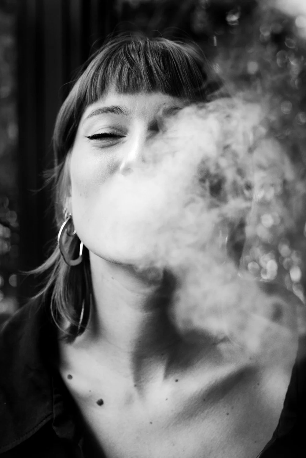 A woman with short hair and hoop earrings is exhaling smoke or vapor, partially obscuring her face.
