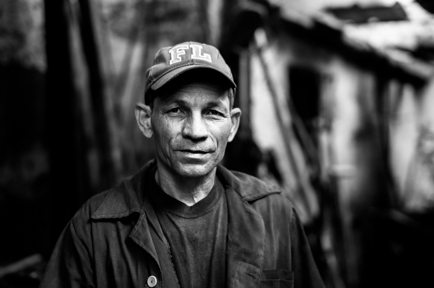 A black and white portrait of a middle-aged man wearing a cap with the letters 'FL' and a jacket, standing indoors with a blurred rustic background.