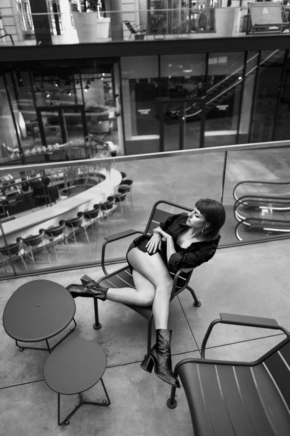 A woman sitting on a chair at an indoor mall or commercial space, dressed in a black dress and boots, with multiple small round tables nearby and escalators behind her.