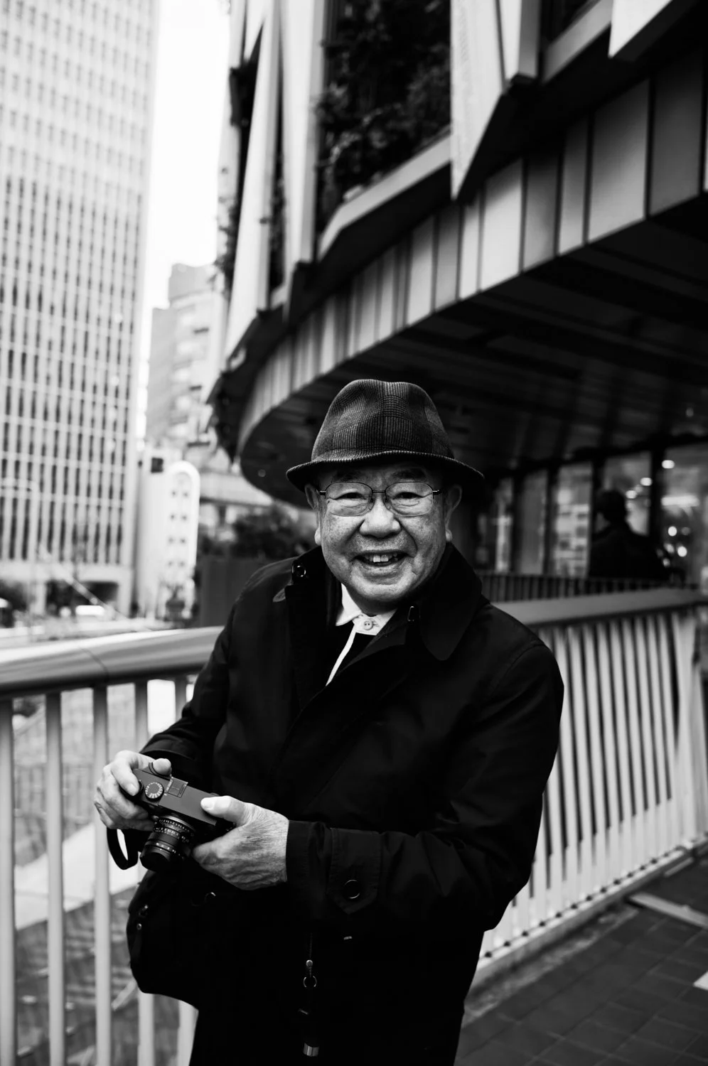 An elderly man smiling and holding a camera on an outdoor balcony in an urban setting, wearing a hat and glasses.