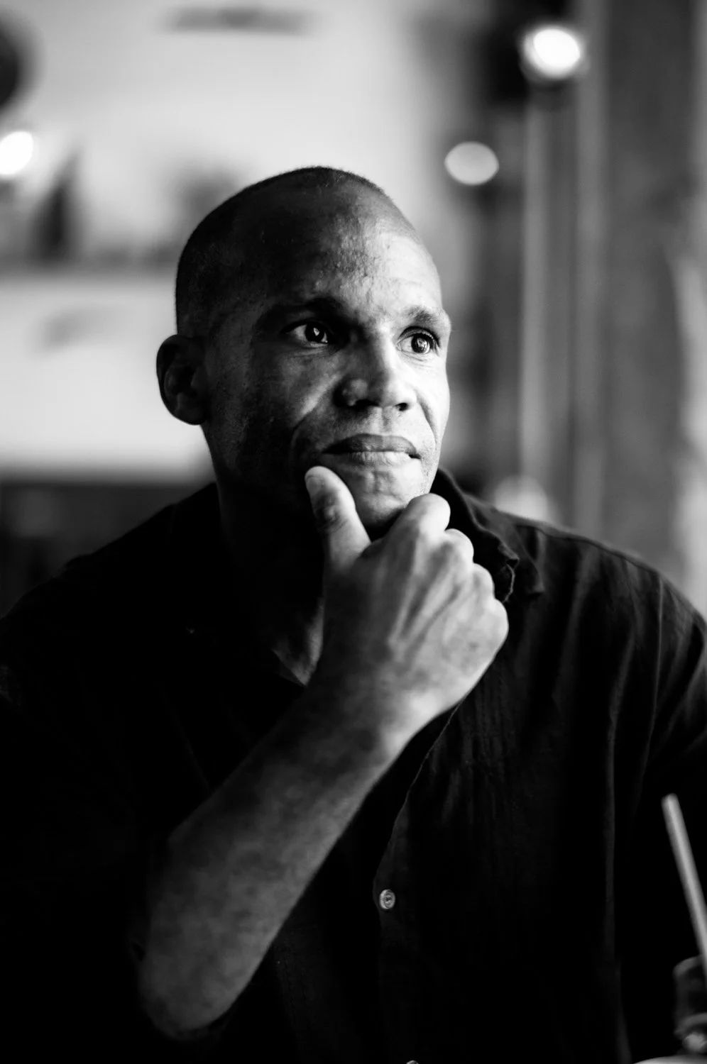 A black and white photo of a man with short hair, sitting indoors, appearing deep in thought with his hand resting on his chin.