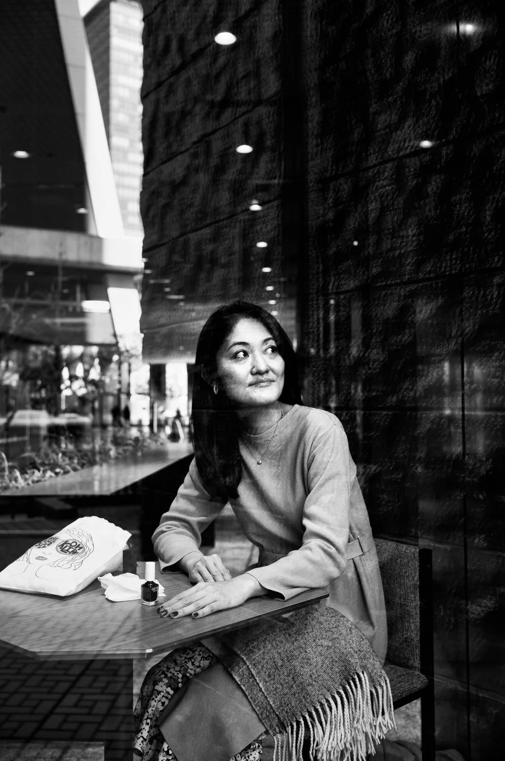 A woman sitting inside a cafe near a window, looking away thoughtfully, with a laptop bag, nail polish, tissues, and a snack on the table.