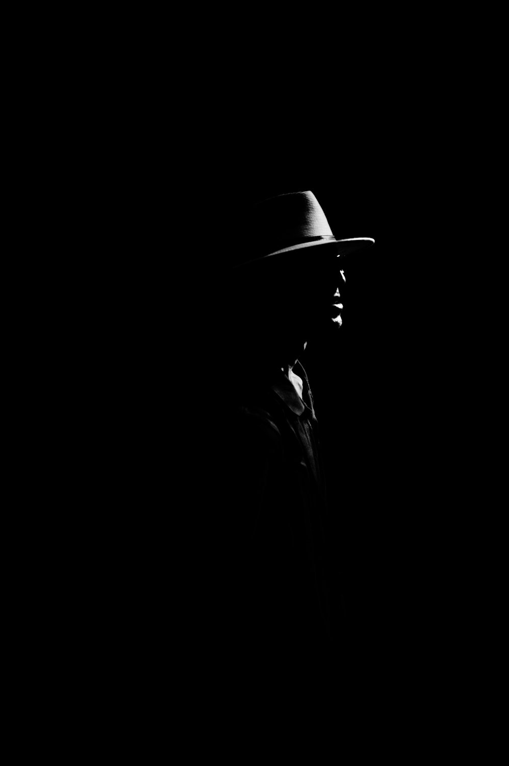 A black and white image of a person wearing a wide-brimmed hat seen in profile, with only part of their face visible against a dark background.