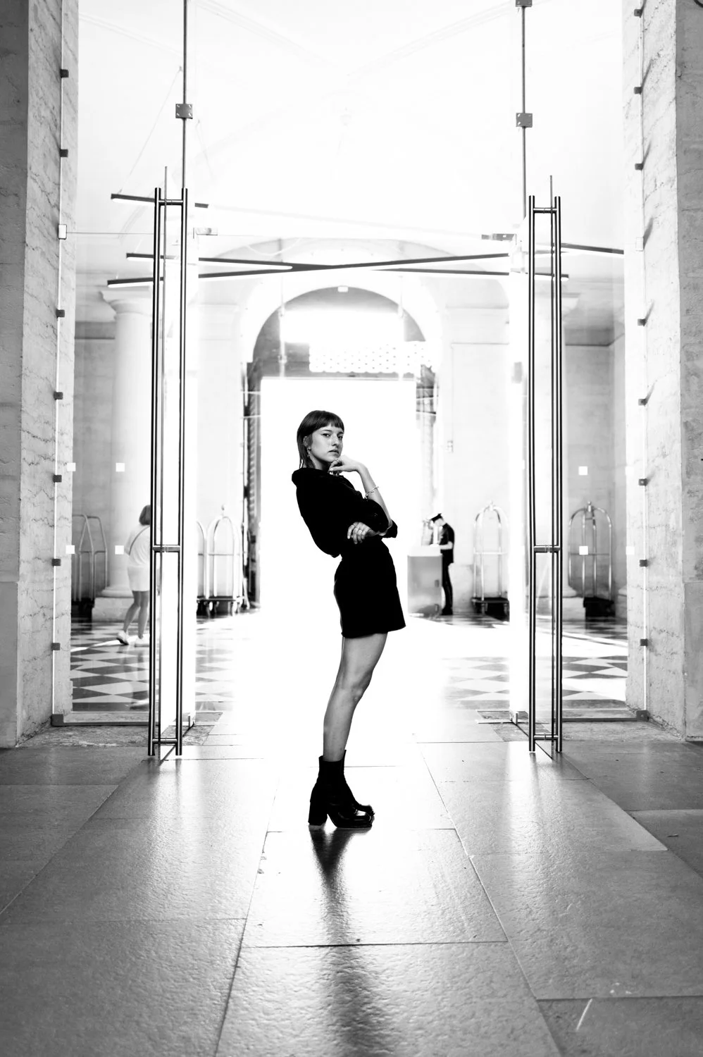 A woman in a black dress and chunky black boots posing inside a large, elegant building with marble pillars and checkered flooring. She is standing in front of a glass door, with her left arm crossed and her right hand touching her chin, looking at t