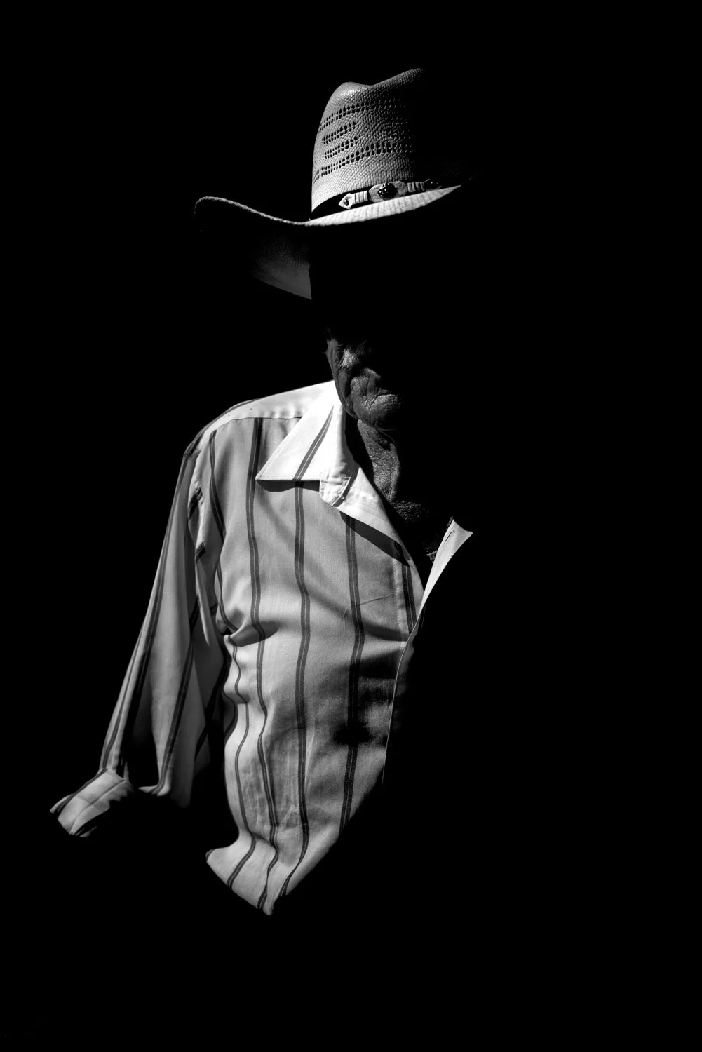 A black-and-white photo of an older man wearing a striped shirt and a cowboy hat, mostly in shadow.
