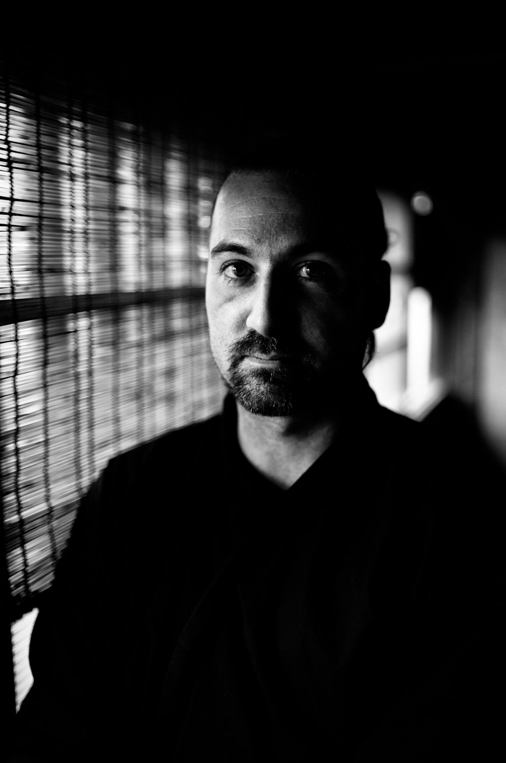 A black-and-white photo of a man with a beard and mustache, looking directly at the camera, standing near a window with blinds, with light coming through the blinds illuminating his face.
