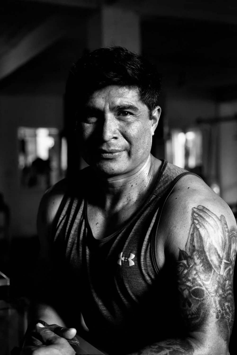 A black-and-white photo of a muscular man with short dark hair, wearing a sleeveless athletic shirt with the Under Armour logo, sitting in a gym.
