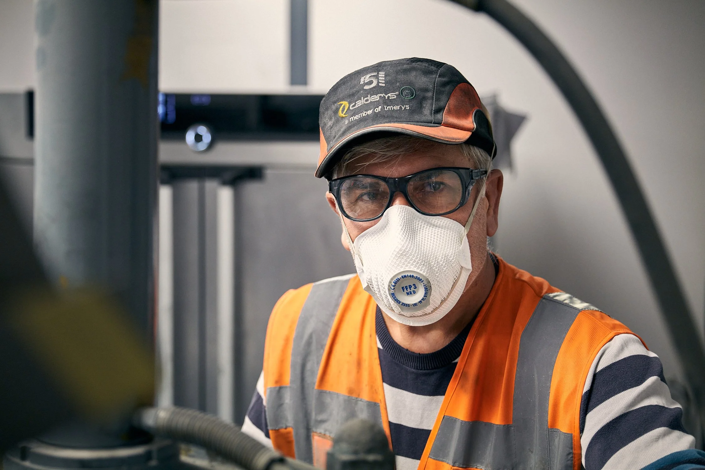 A man wearing safety glasses, a face mask with an FFP3 label, an orange safety vest, and a cap, working in an industrial or construction setting.