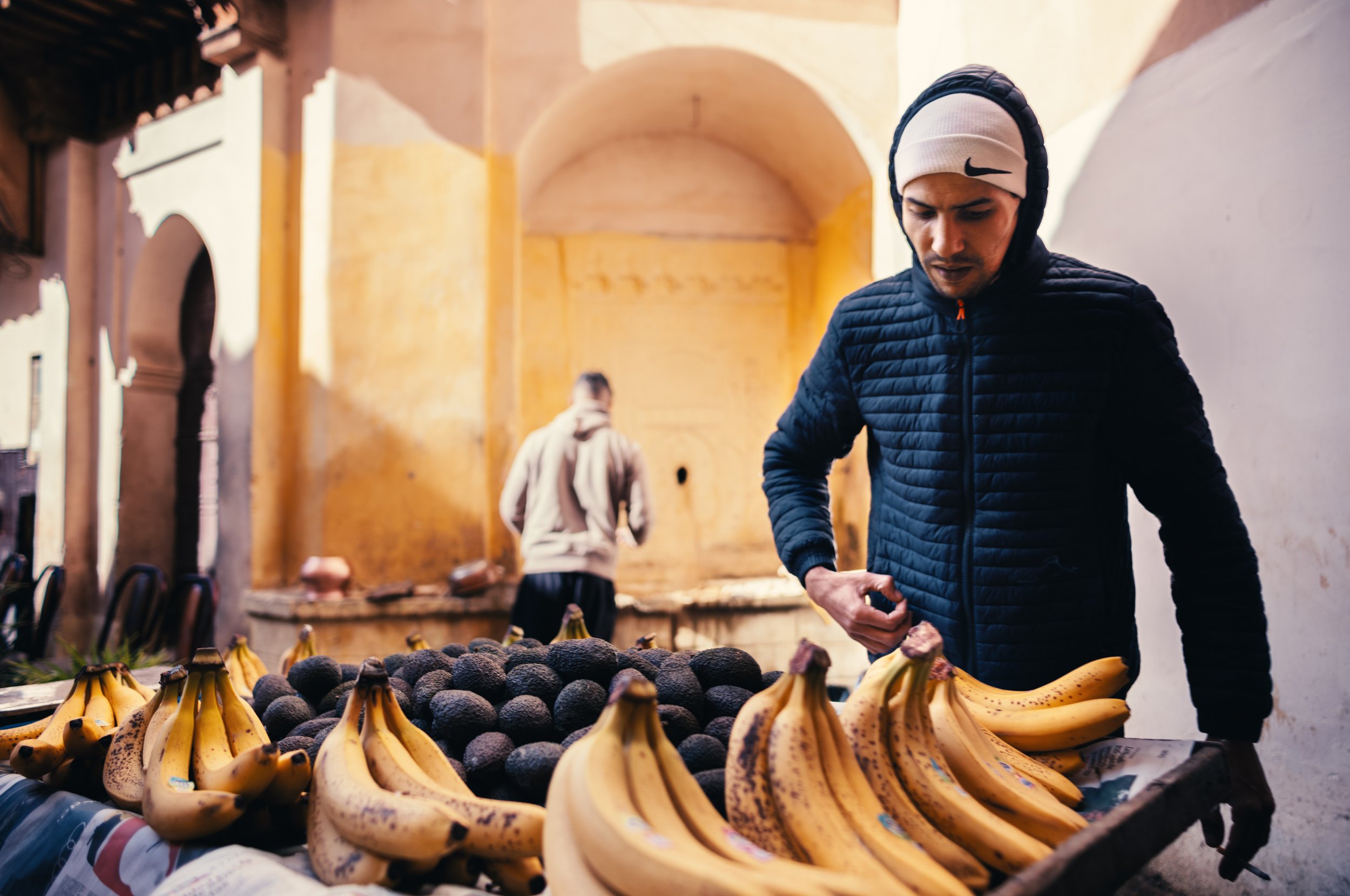 Man shopping for bananas and avocados at a street market with colorful, rustic walls in the background.