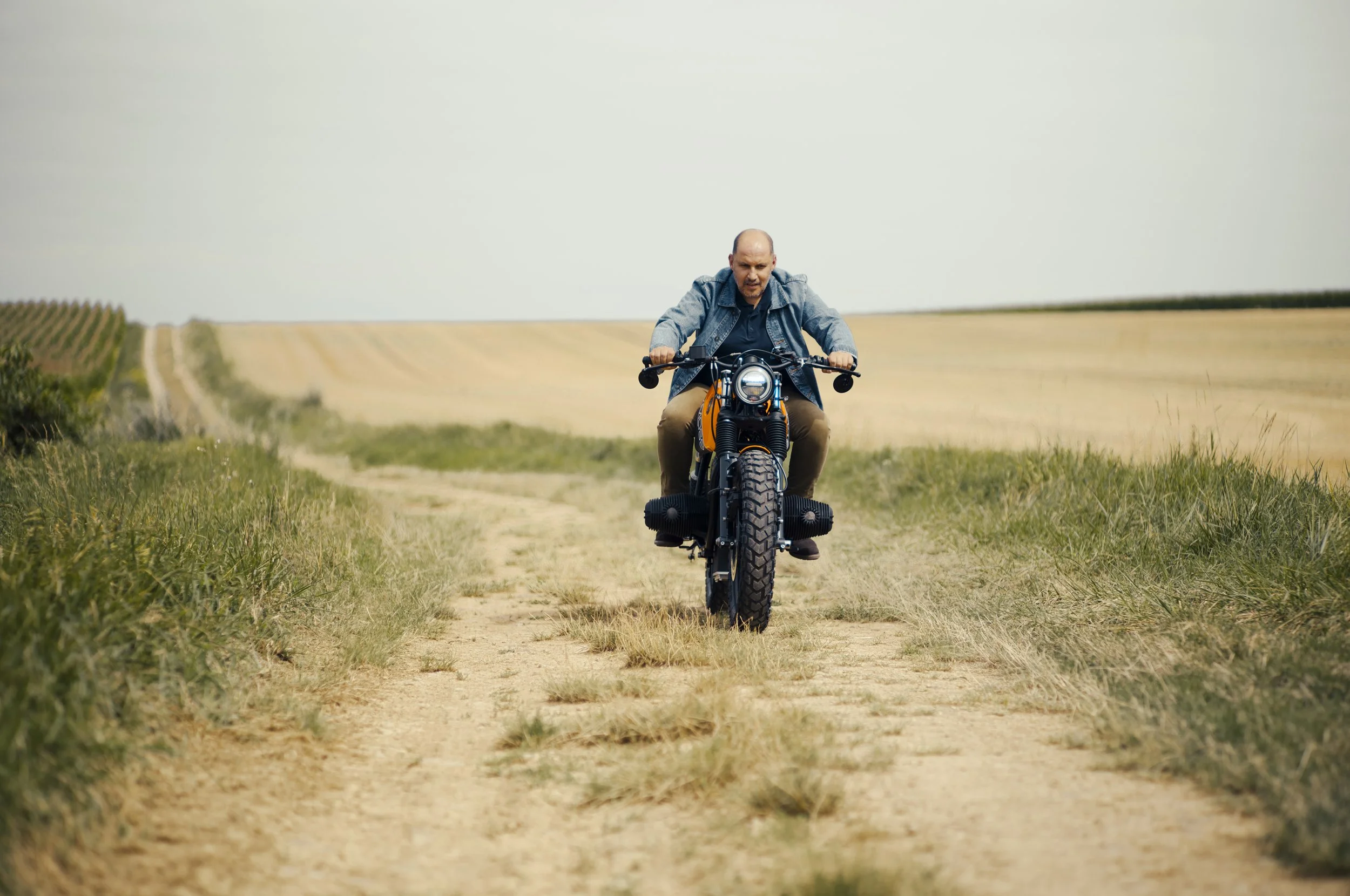 A man riding a motorcycle on a dirt path through a rural field under an overcast sky.