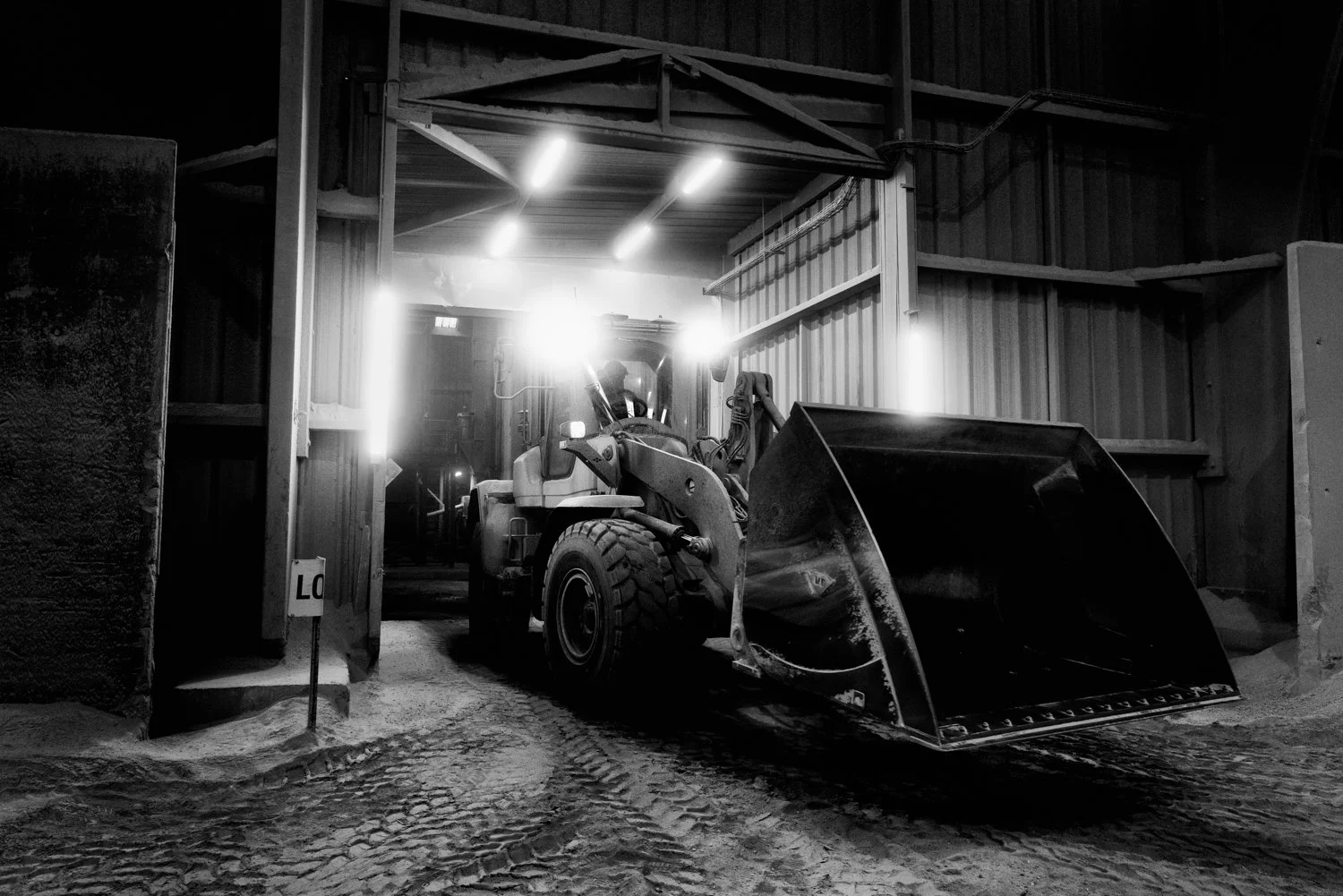 A bulldozer inside a warehouse or garage with bright lights overhead, with some snow or dirt on the ground outside the entrance.