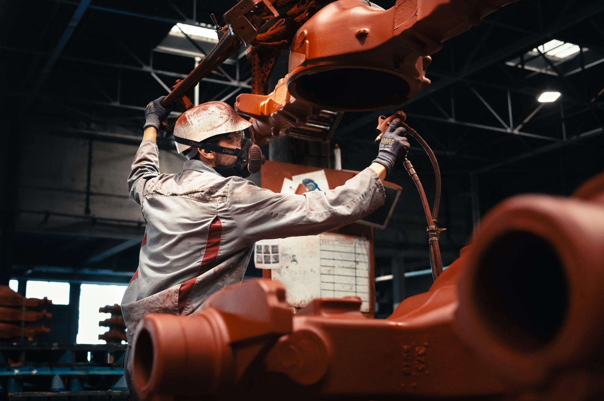 Worker wearing safety gear and a helmet, performing welding or maintenance on a large industrial machine in a factory or workshop.