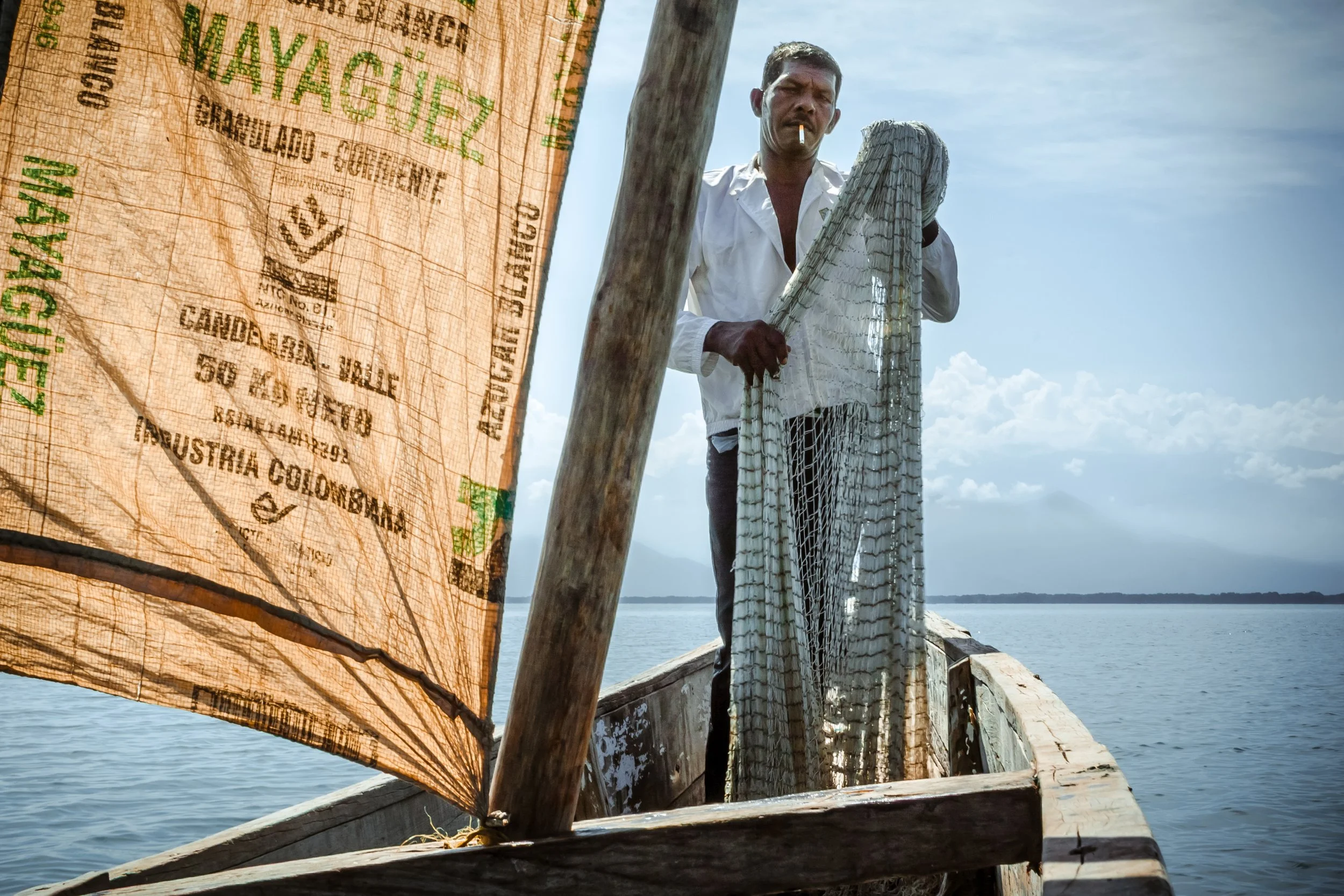 A man standing on a wooden boat holding a fishing net, with a river and sky in the background, and a cloth with text in the foreground.