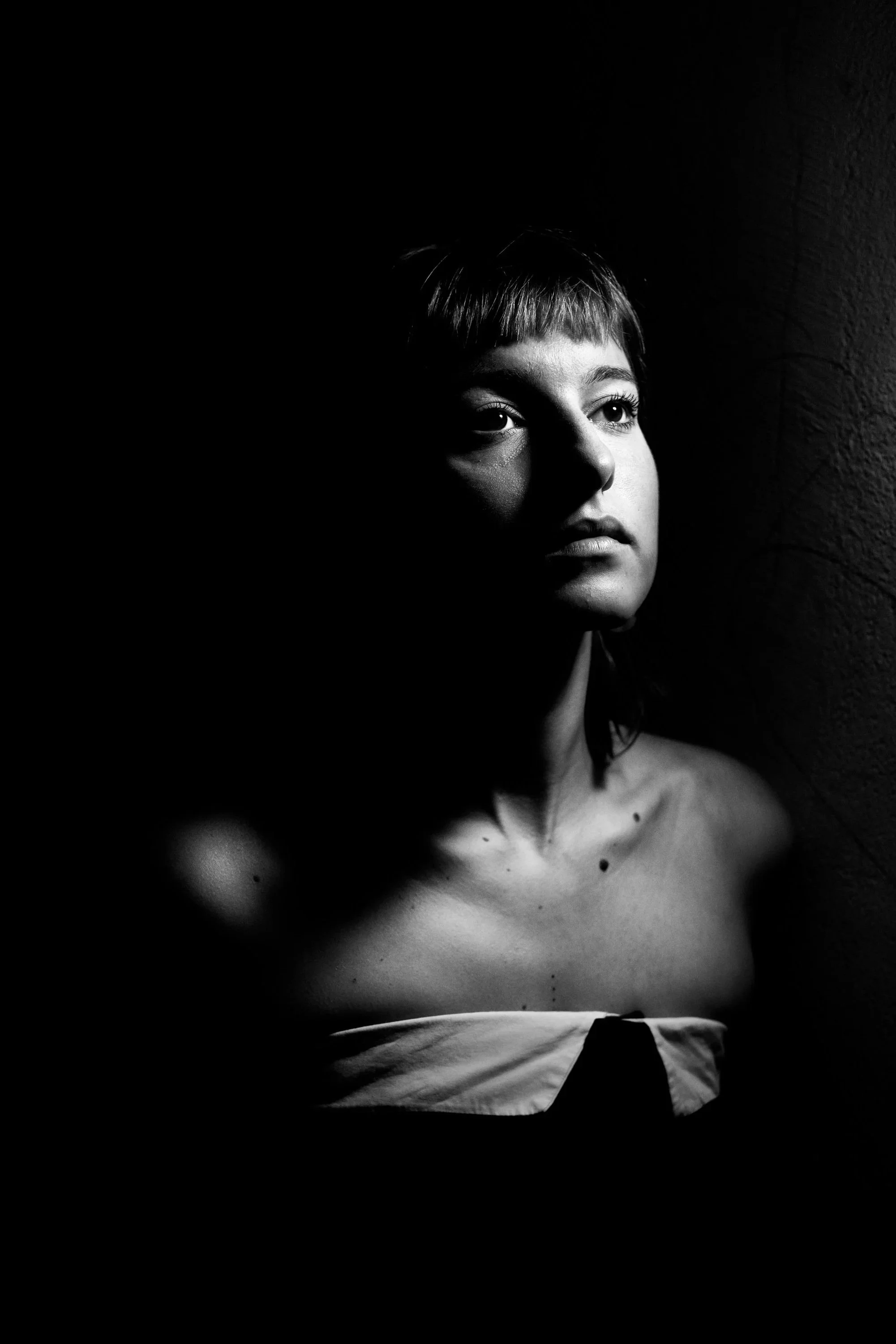 Black-and-white portrait of a young woman with short hair, illuminated by a single light source from the right, creating shadows on her face and shoulder, with a textured wall in the background.