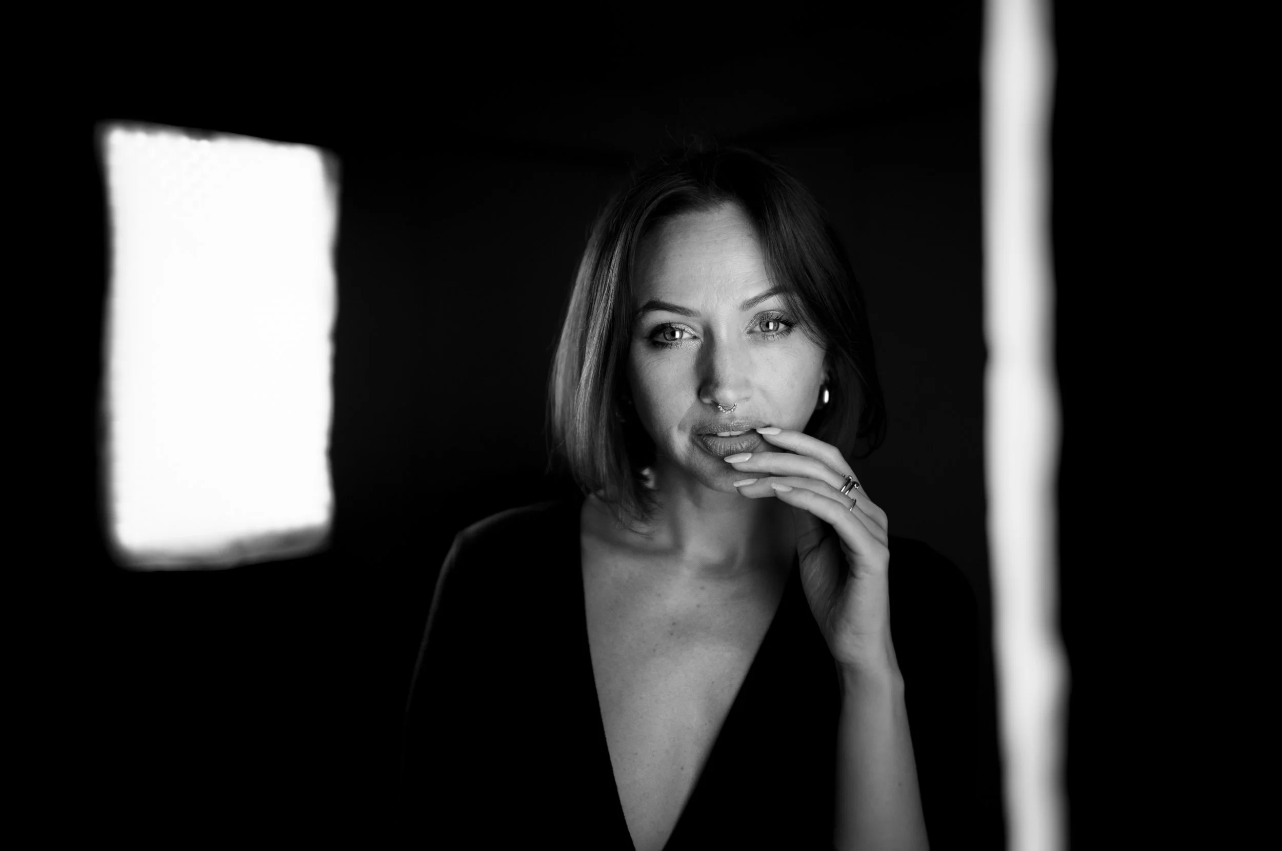 Black and white photo of a woman with short hair, gazing thoughtfully at the camera, with her hand near her mouth. The photo appears to be taken through a mirror or frame.