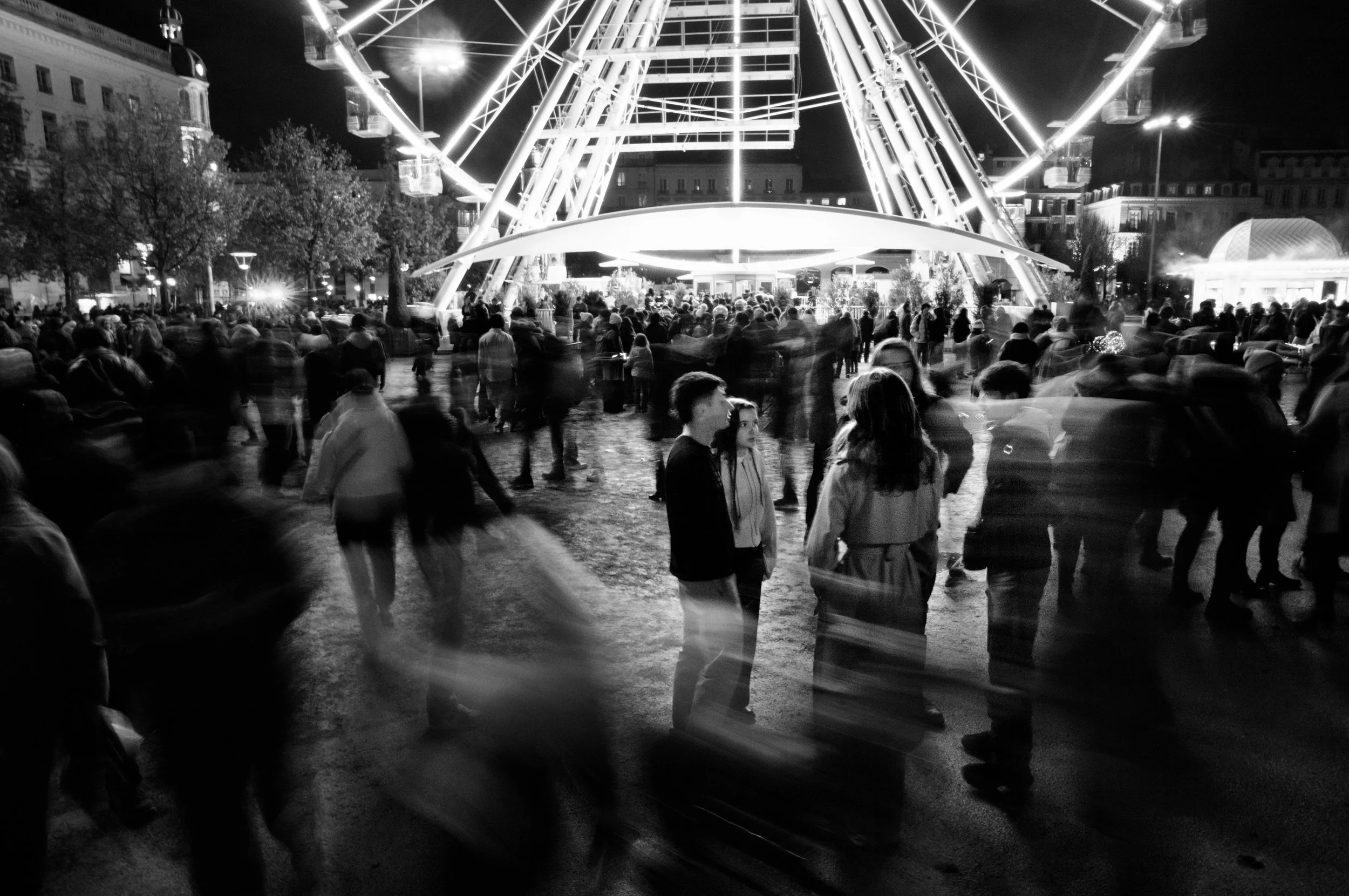 Night scene of a crowded outdoor city fair with a large illuminated Ferris wheel in the background, where numerous people are walking and standing around, some appearing blurred due to motion.
