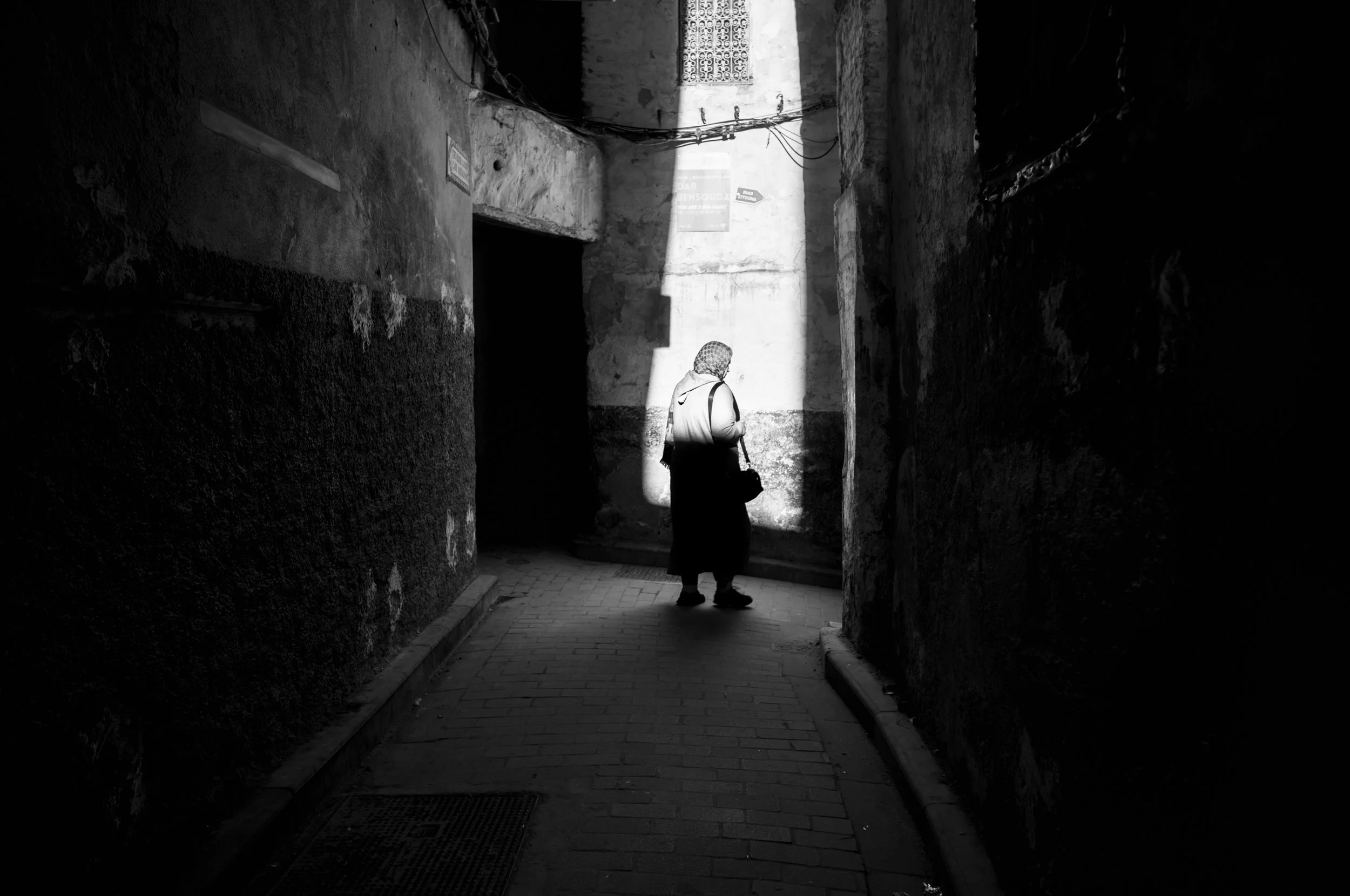 A person standing in a narrow, dark alleyway, illuminated by a beam of sunlight, with textured walls and overhead electrical wires.
