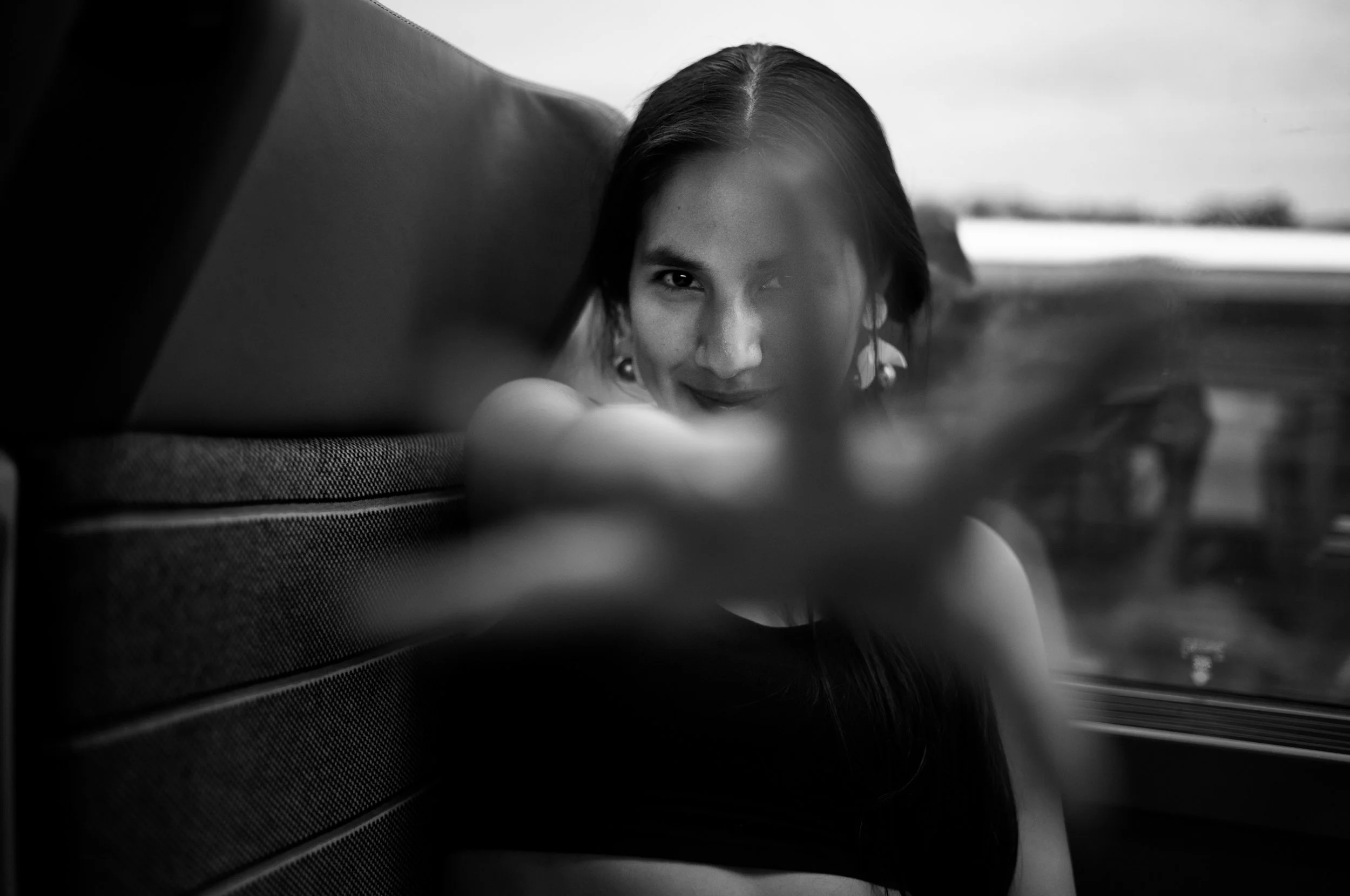 Black and white photo of a woman sitting in a vehicle, leaning forward with her face partially obscured by her hand, wearing earrings, with a blurred view outside the window.