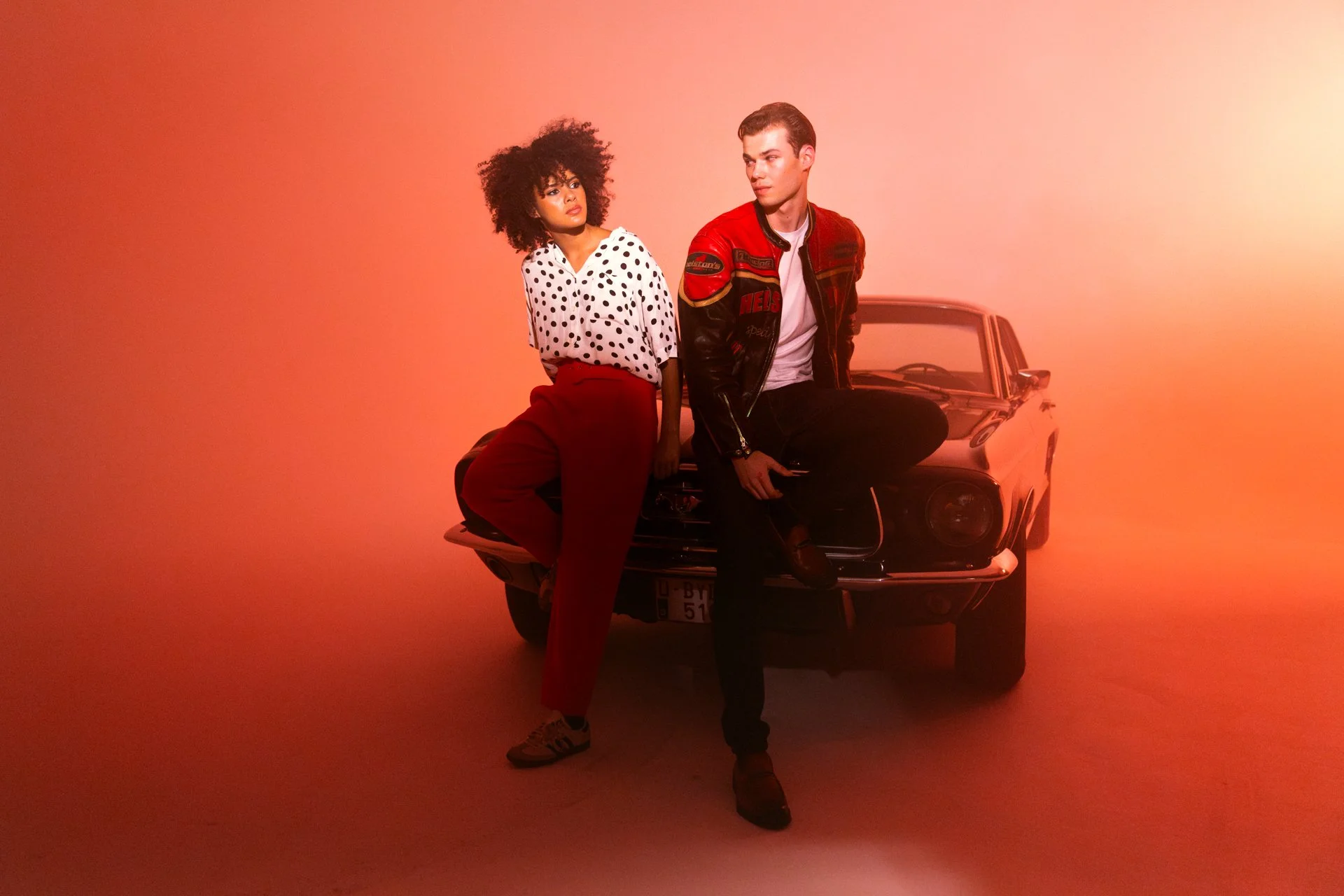 A woman with curly hair and a man with short hair sitting on the front of a classic black car in a studio with a warm orange background.