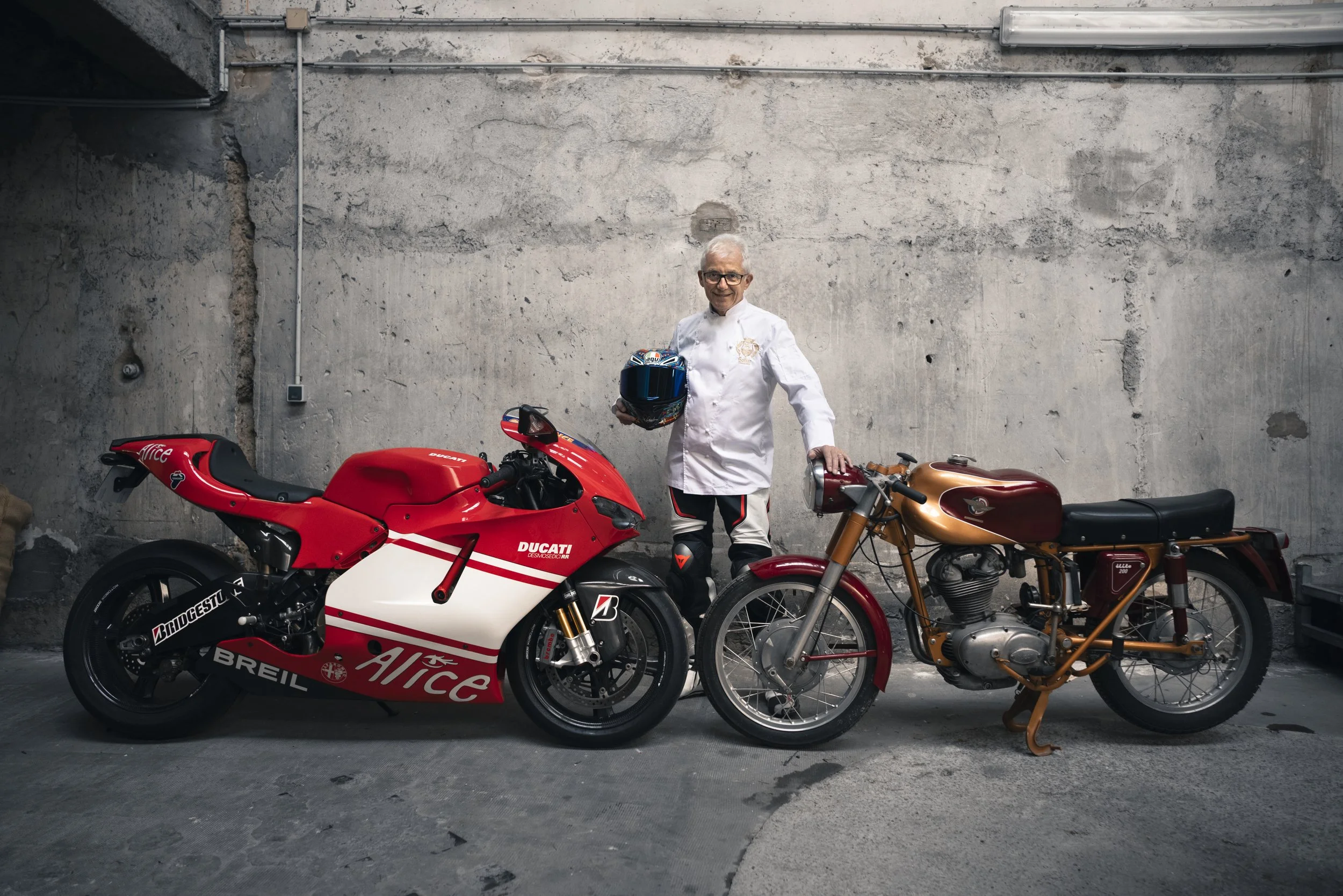 Older man in a white racing suit standing between two vintage motorcycles in a garage