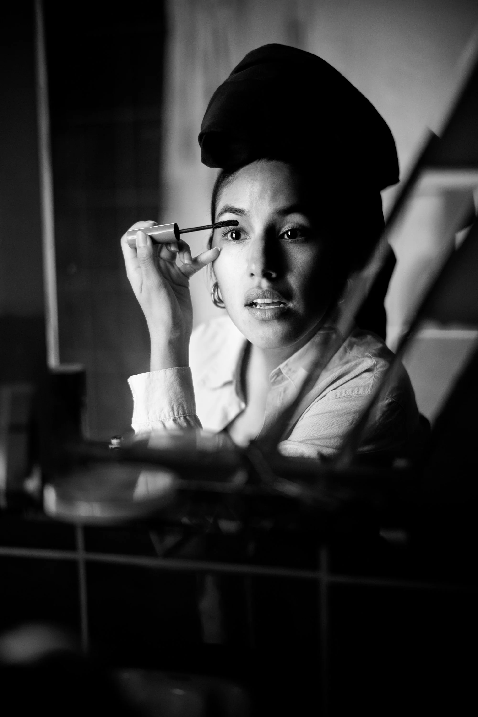 A woman applying mascara in front of a mirror, with a towel wrapped around her head, in black and white.