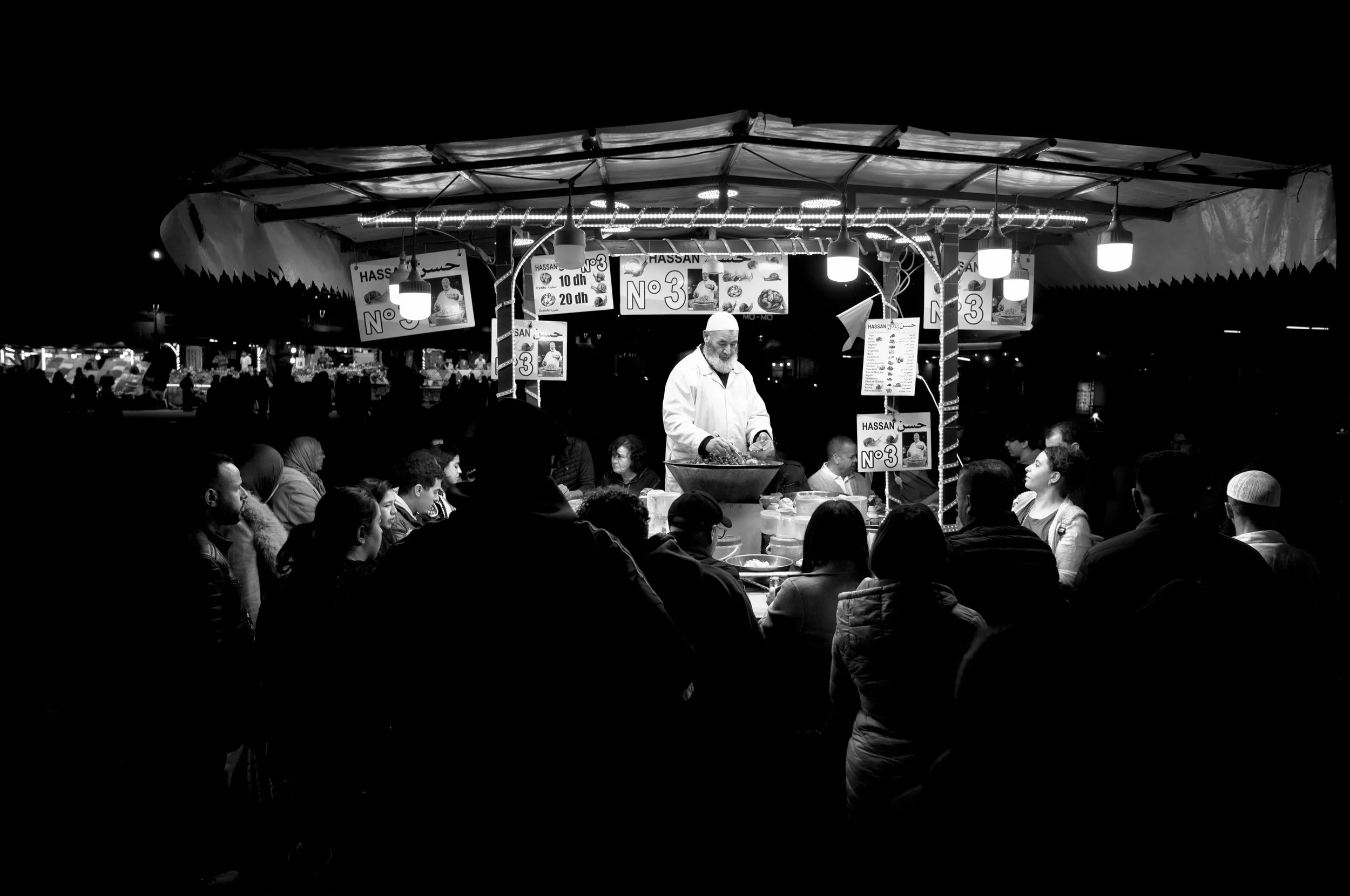 A street food stall at night with a crowd of people gathered around it. The vendor is preparing food under a canopy with illuminated signs in Arabic and English, advertising their offerings and prices. The scene is in black and white.
