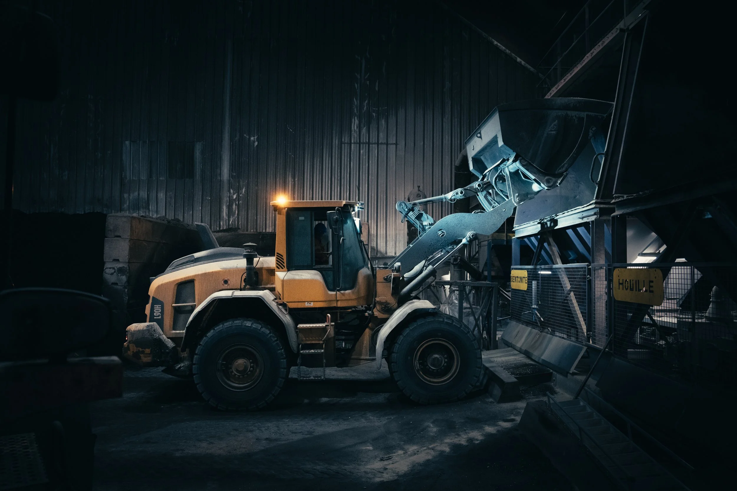 A yellow construction vehicle shovel loader inside a dimly lit industrial warehouse, with piles of materials and construction equipment around.