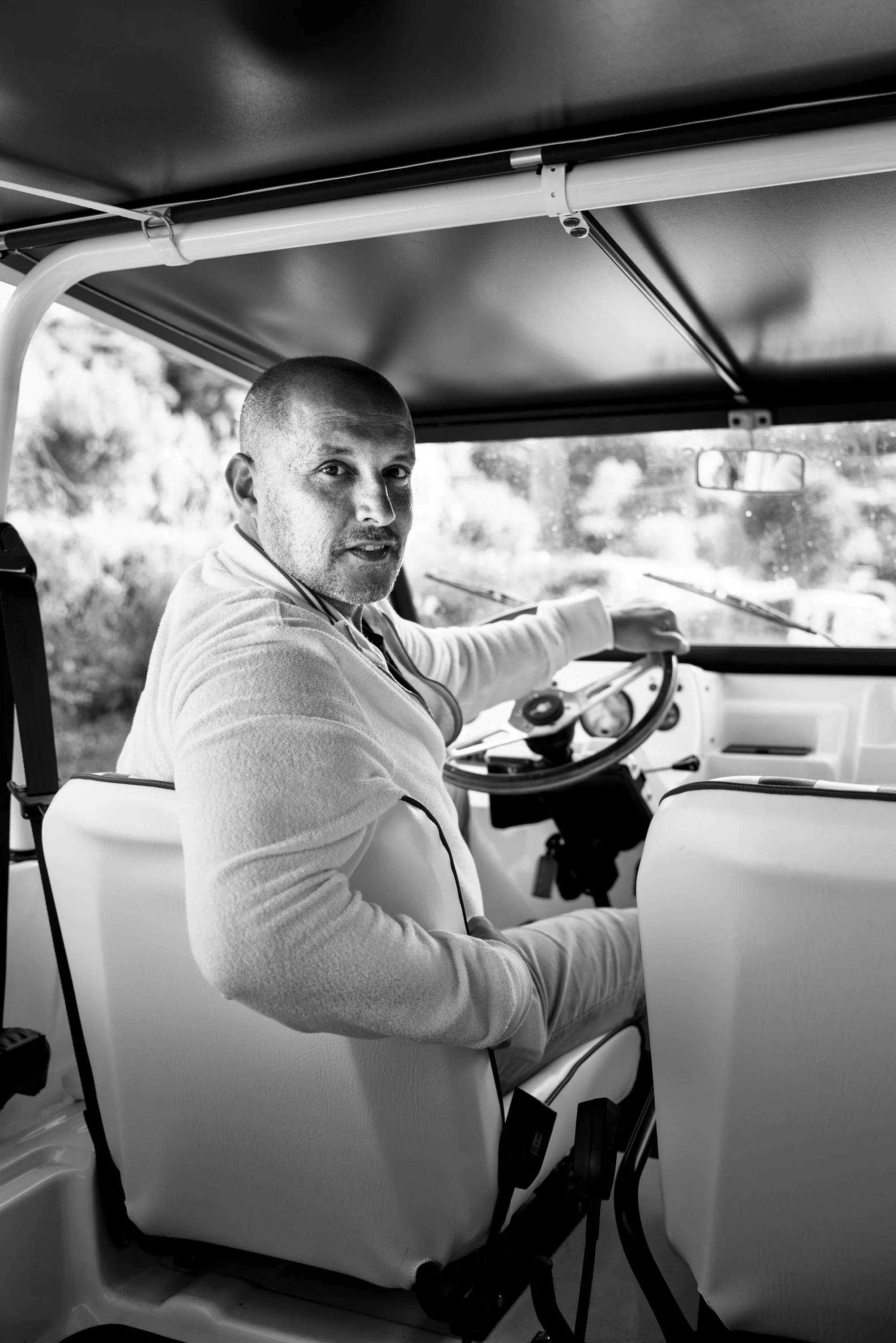 Man with a shaved head sitting in the driver's seat of a vehicle, looking over his shoulder, with trees and the road visible through the windshield.