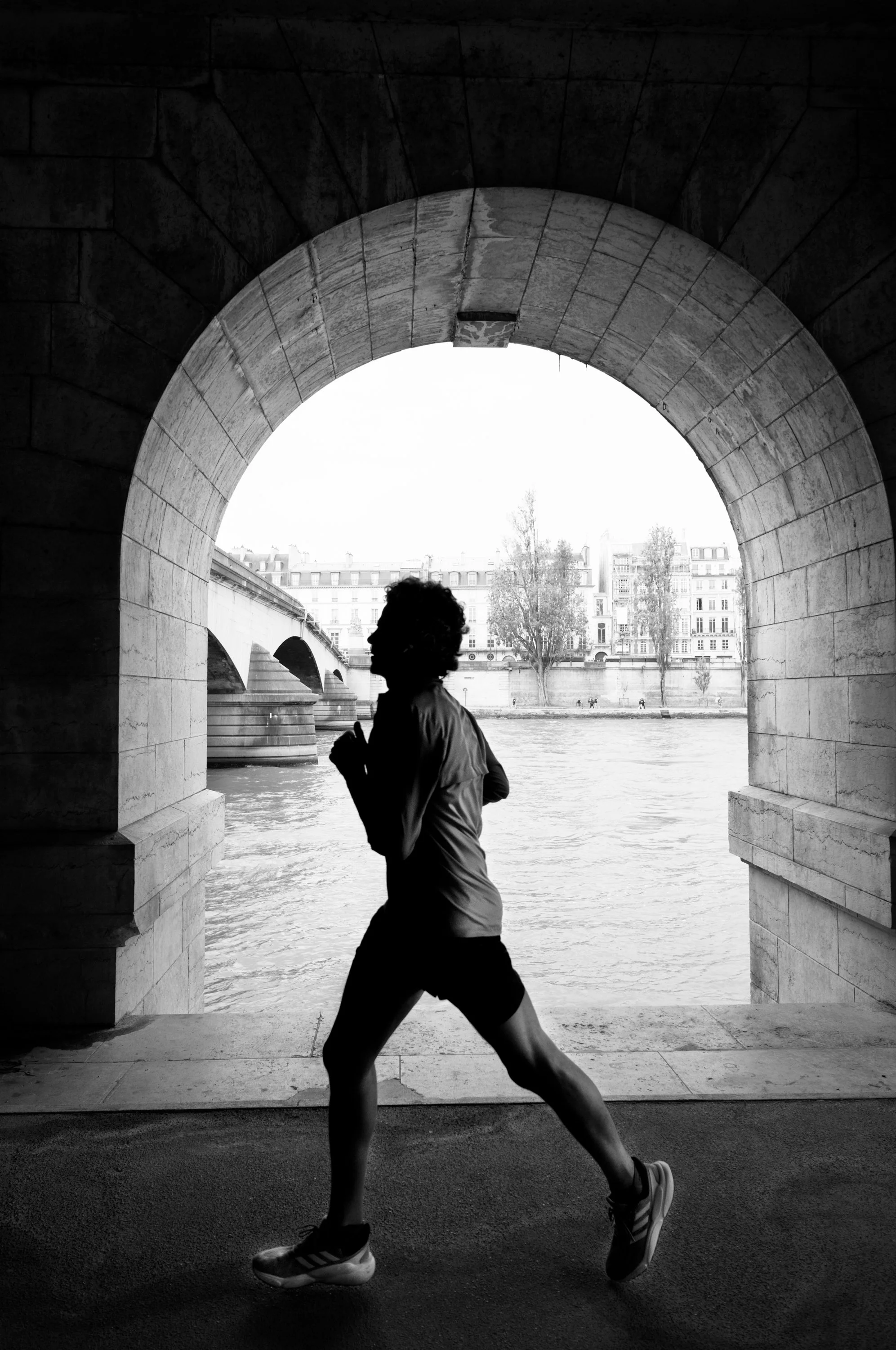 A person running under a stone archway near a river with buildings and trees in the background in black and white.