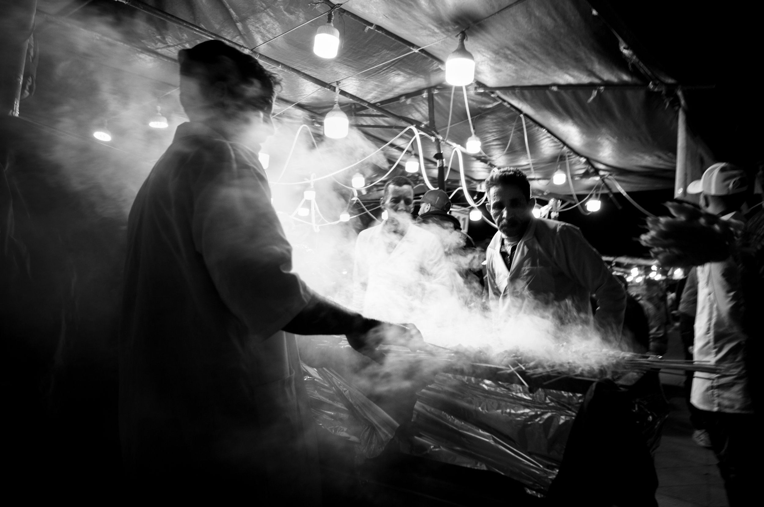 People working at a food stall with steam rising, under a tent with hanging lights at night.