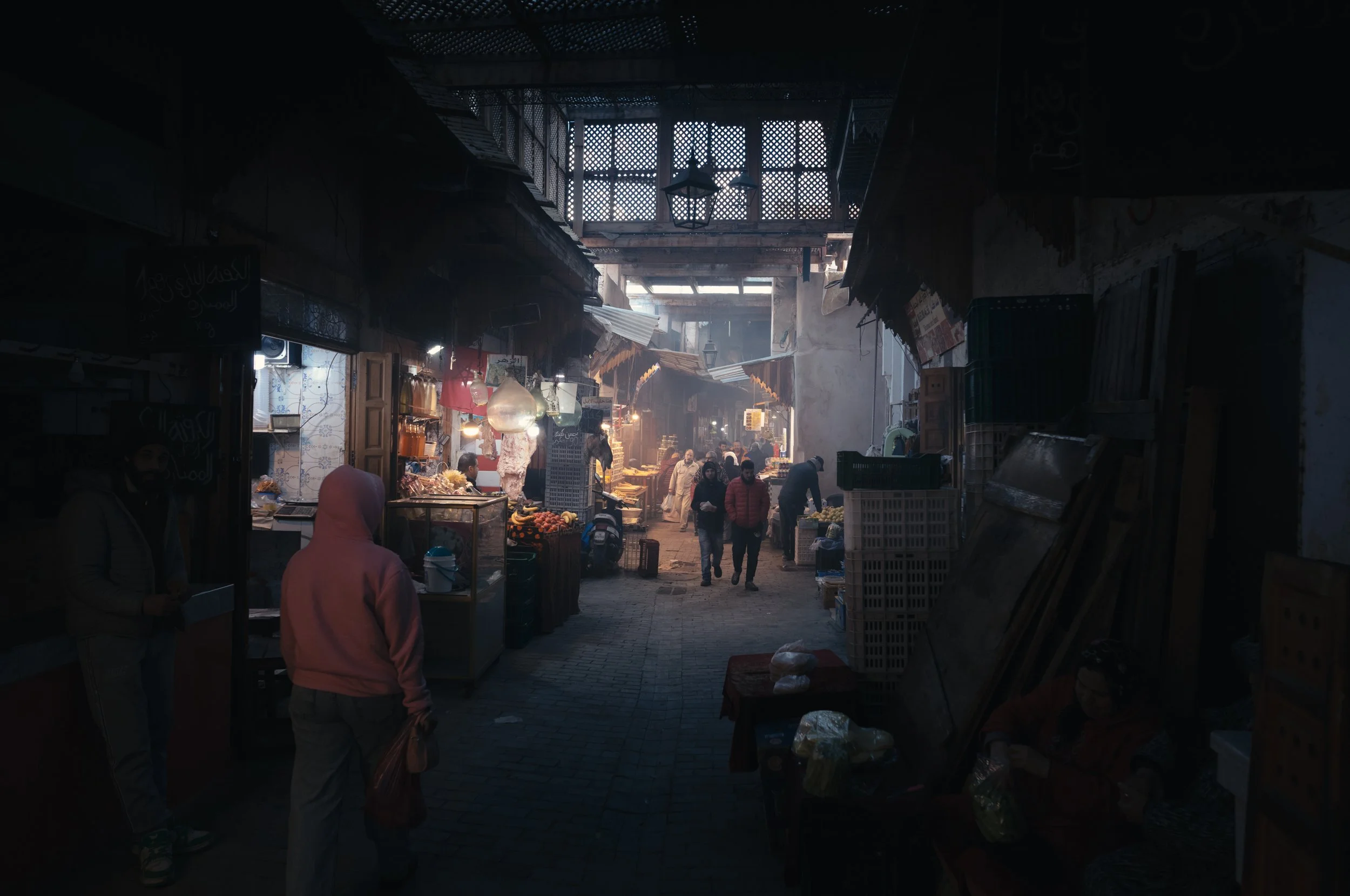 A busy indoor market with various food stalls and vendors, people walking and shopping, sunlight streaming through a silver patterned upper window.