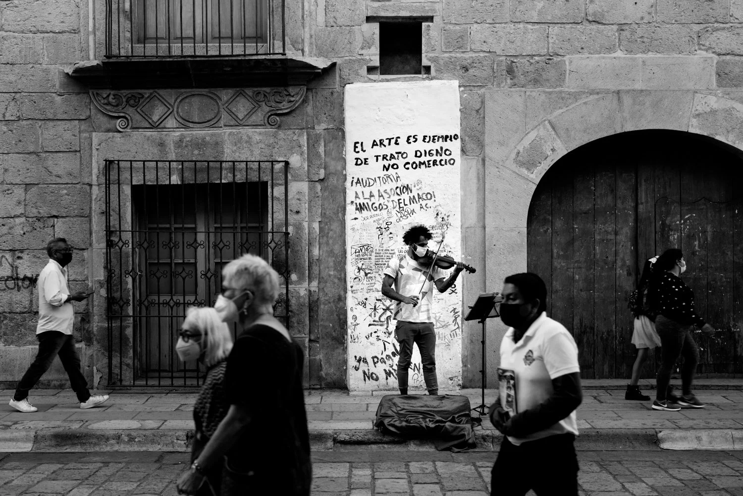 Street scene with a violin player performing on a sidewalk in front of a graffiti-covered wall, surrounded by pedestrians wearing masks.