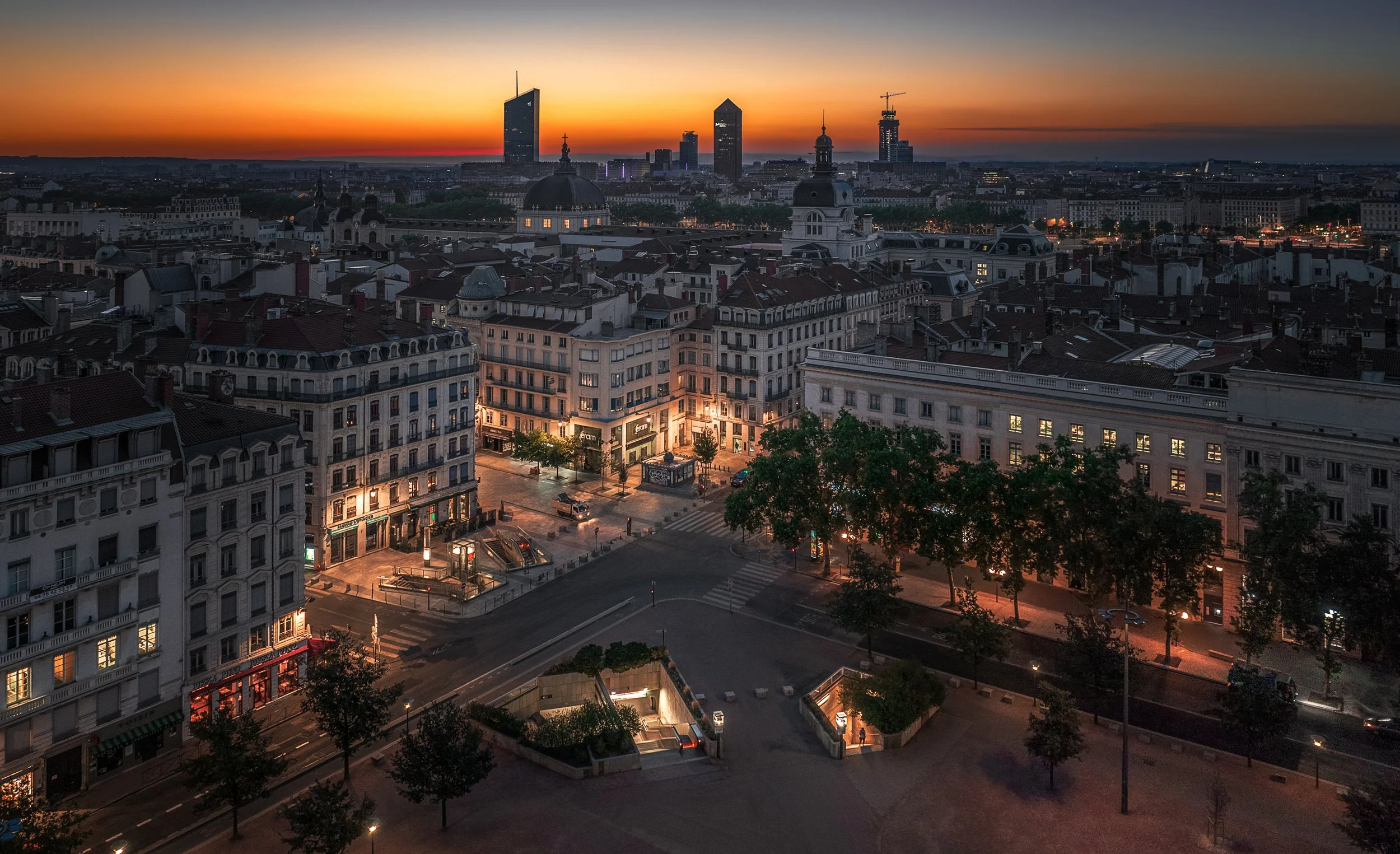 City skyline at sunset with tall buildings, some under construction, historic architecture, and streets illuminated by streetlights.