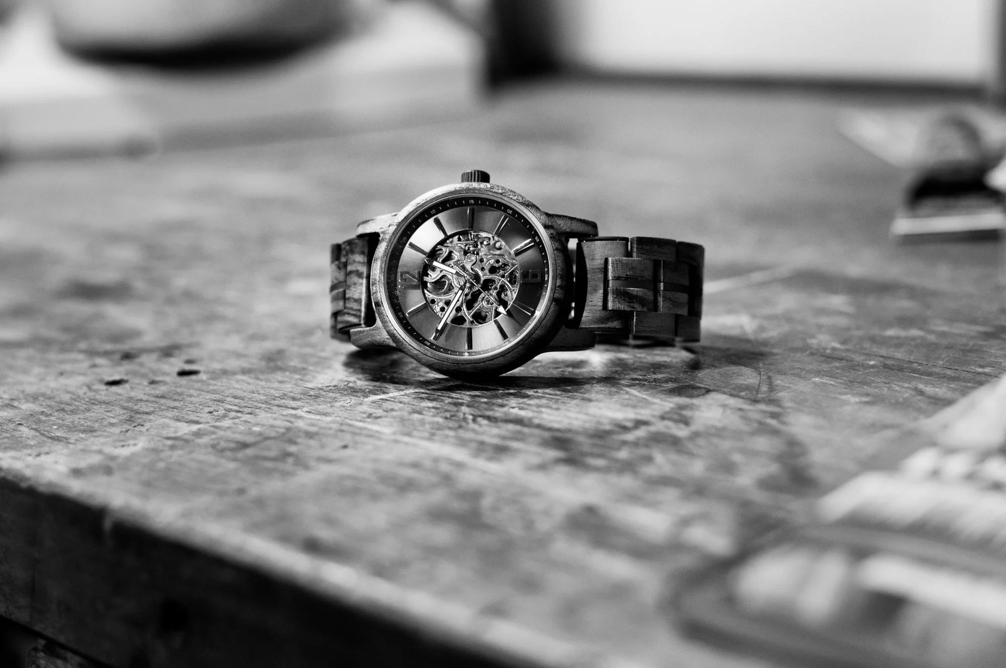 A metal wristwatch with a visible mechanical movement lies on a wooden surface in black and white.