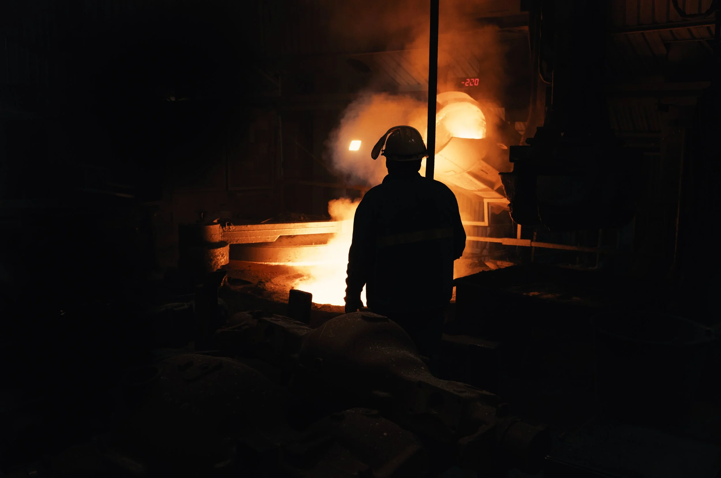 A firefighter silhouette standing in front of a large industrial furnace or molten metal pool with bright orange glow and smoke, inside a factory or foundry.