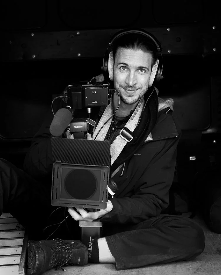 Man sitting on the floor with headphones, holding a camera and camera filter, smiling at the camera.