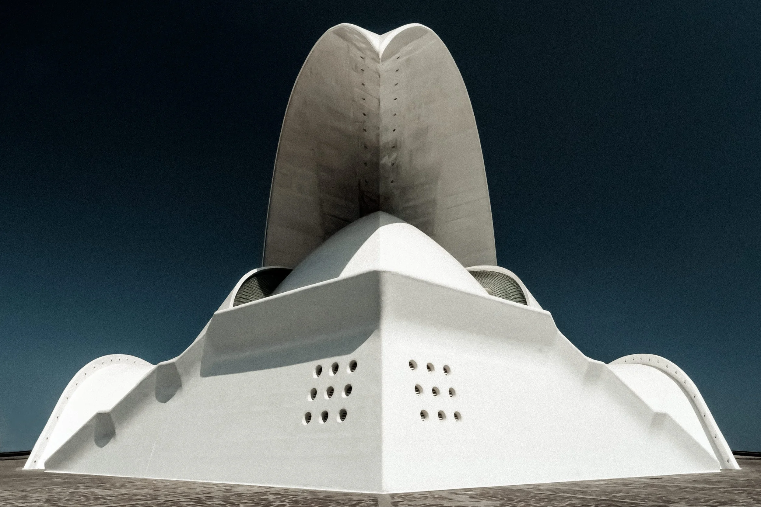 Low-angle view of a modern architectural building with white curved surfaces and large vertical concrete panels against a dark blue sky.
