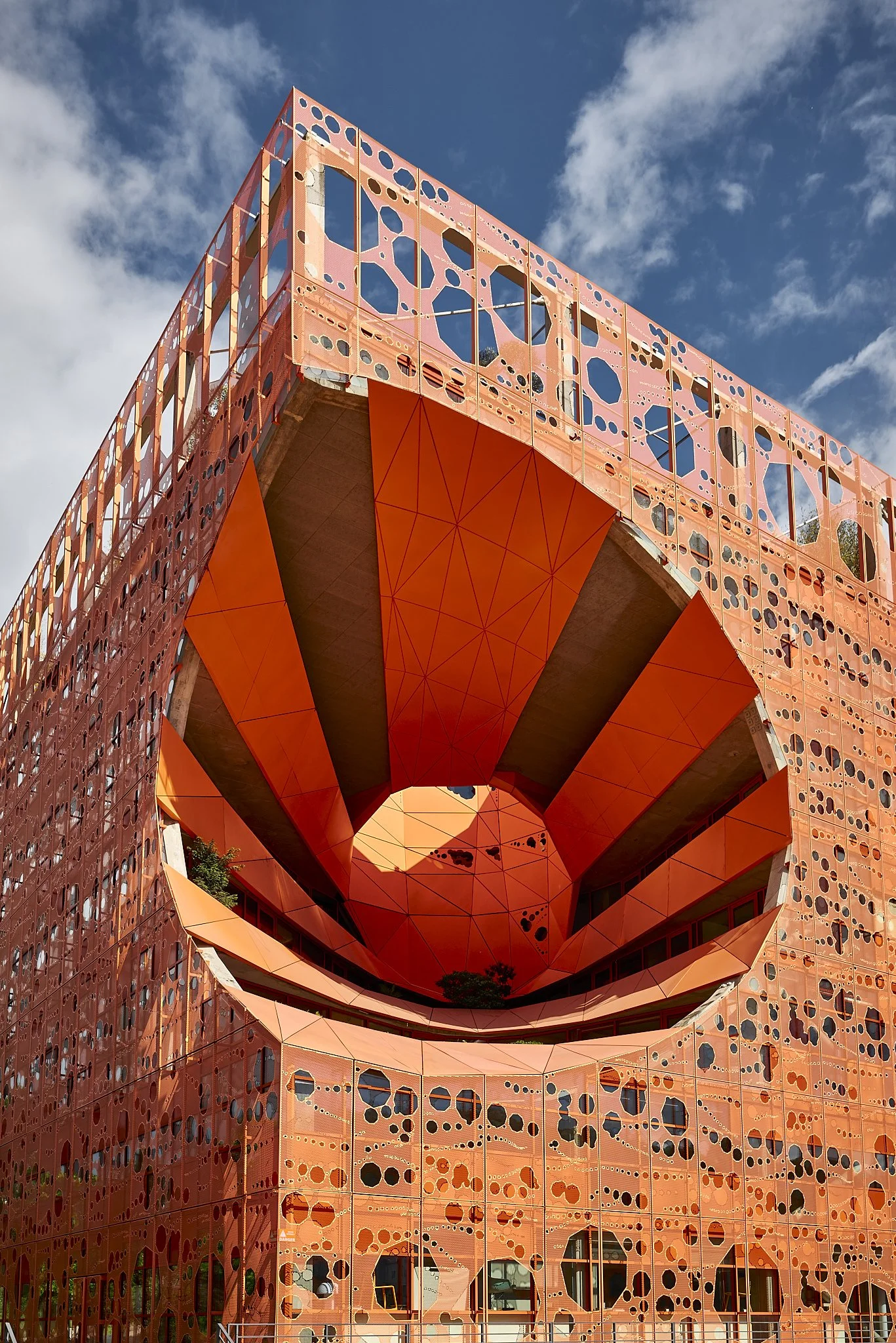 A modern architectural building with a large central hole, orange panels, and patterned facade against a blue sky with clouds.