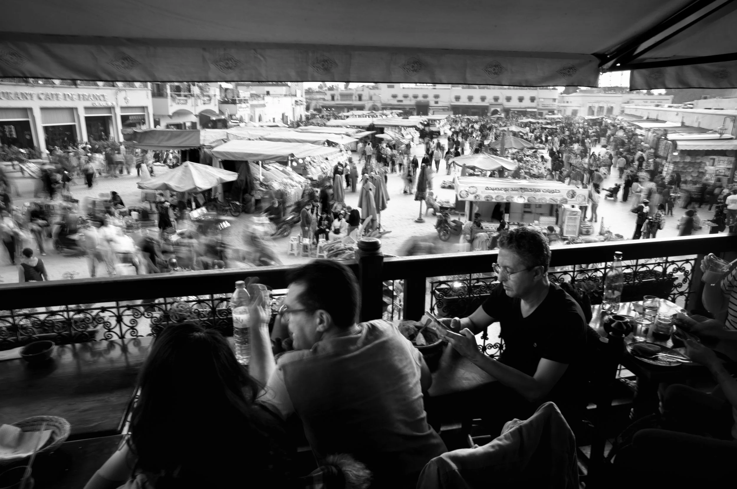 View from a restaurant balcony overlooking a busy marketplace with numerous stalls and people walking, in black and white.
