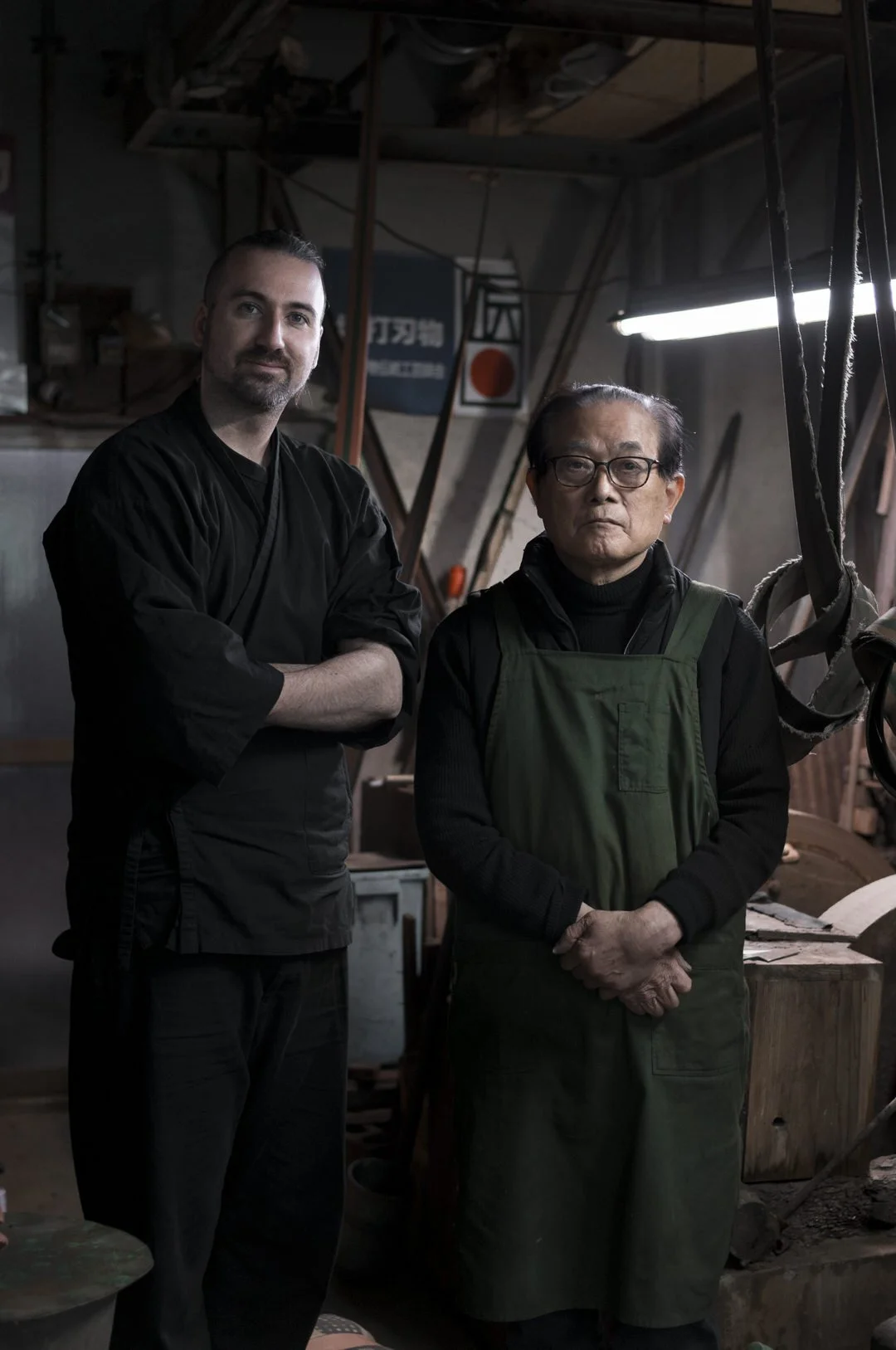 Two men standing in a workshop or woodworking space, wearing black and green aprons, with tools and wood around them.
