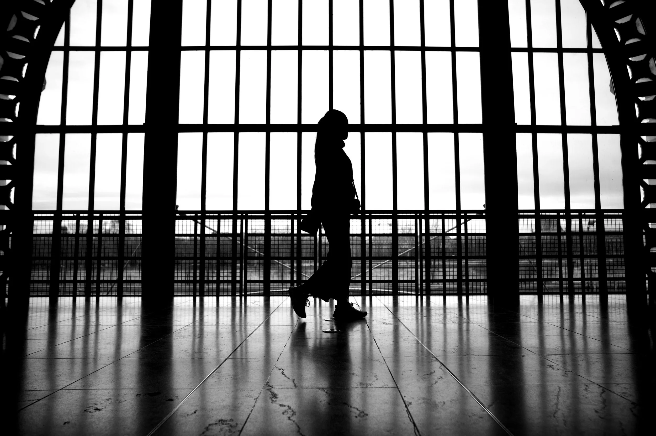 Silhouette of a woman in ice skates standing inside a building with large windows and a view of a body of water outside.