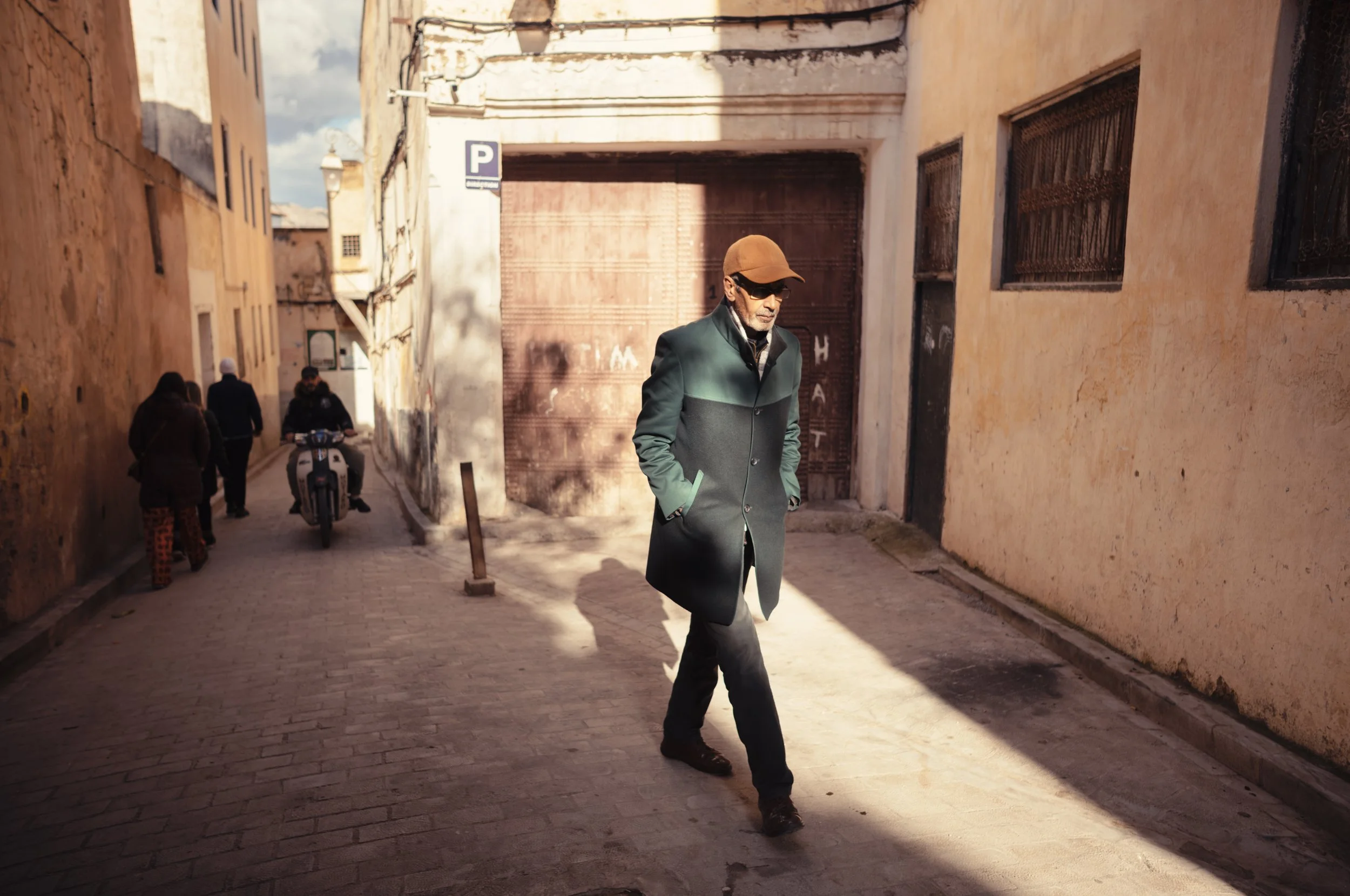 A man wearing a brown cap, sunglasses, and a long dark green coat walking on a cobblestone street lined with yellowish buildings. Other pedestrians and a person on a scooter are visible in the background.