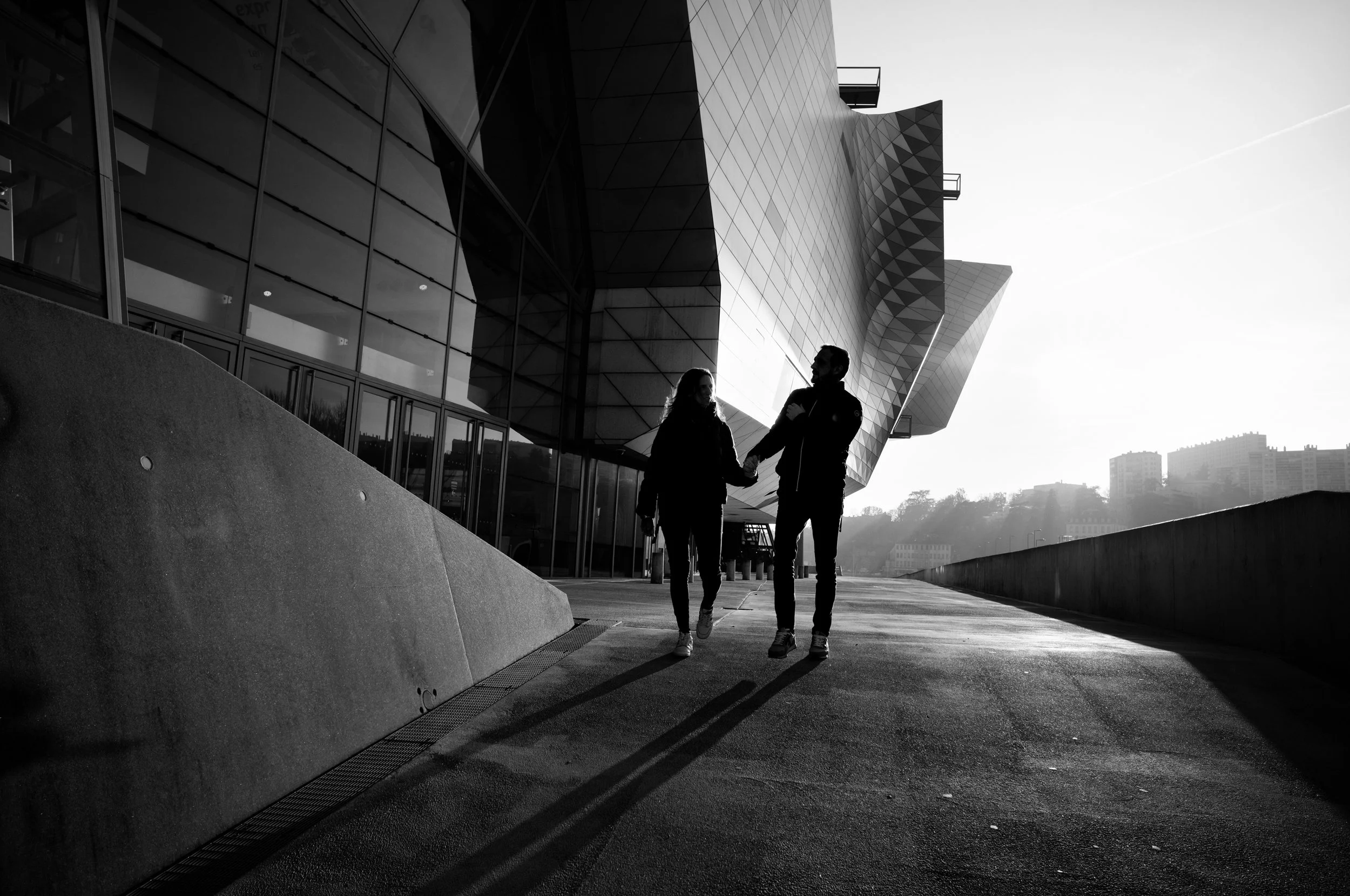 Silhouettes of a man and woman walking and holding hands outside a modern building with geometric architecture, during sunset, in black and white.